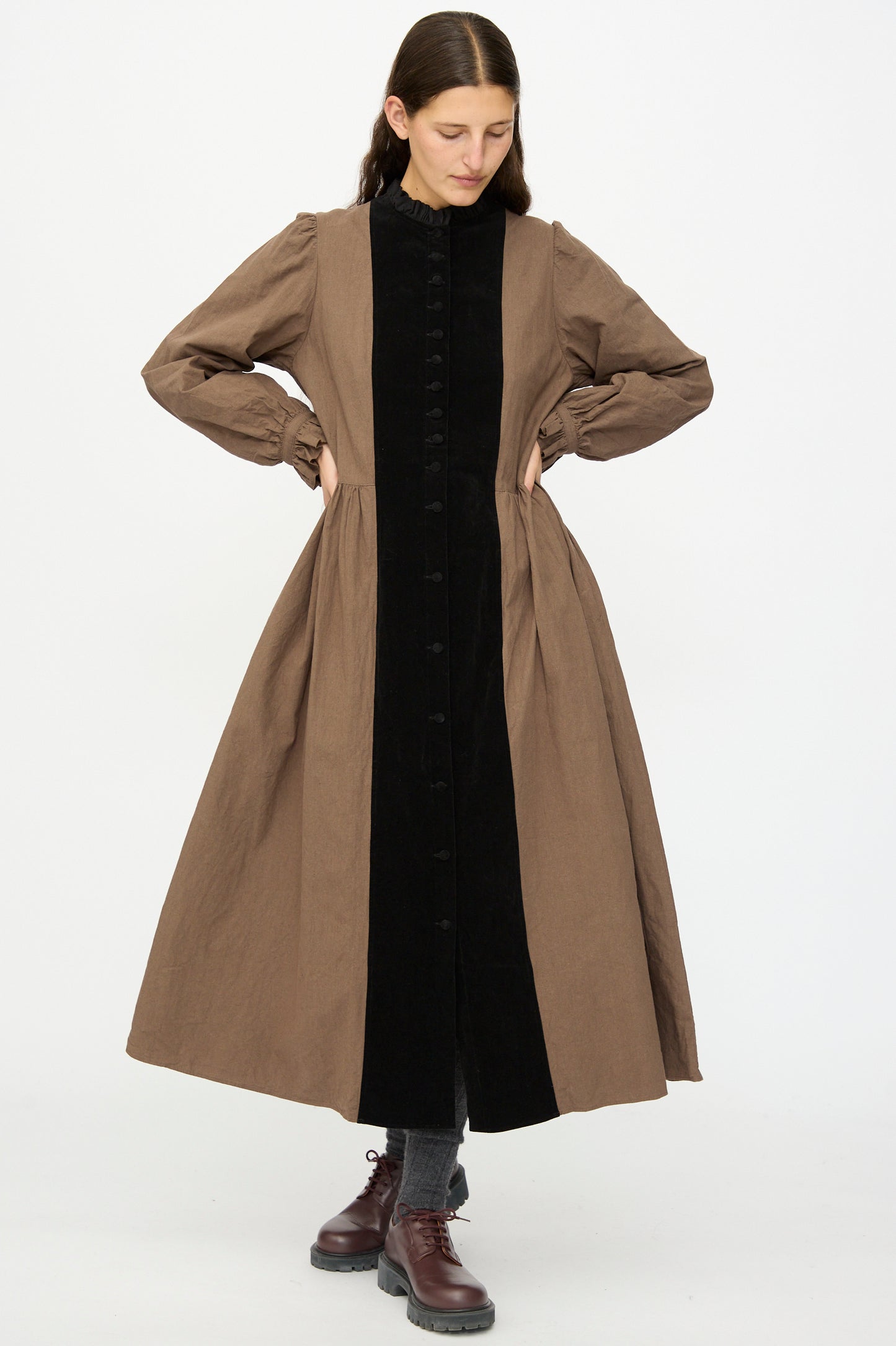 A person stands against a white background wearing Hallelujah’s Jeune Paysanne in Chocolate and Black, a long cotton dress made in Japan, paired with gray socks and brown lace-up shoes—an ensemble of understated elegance.