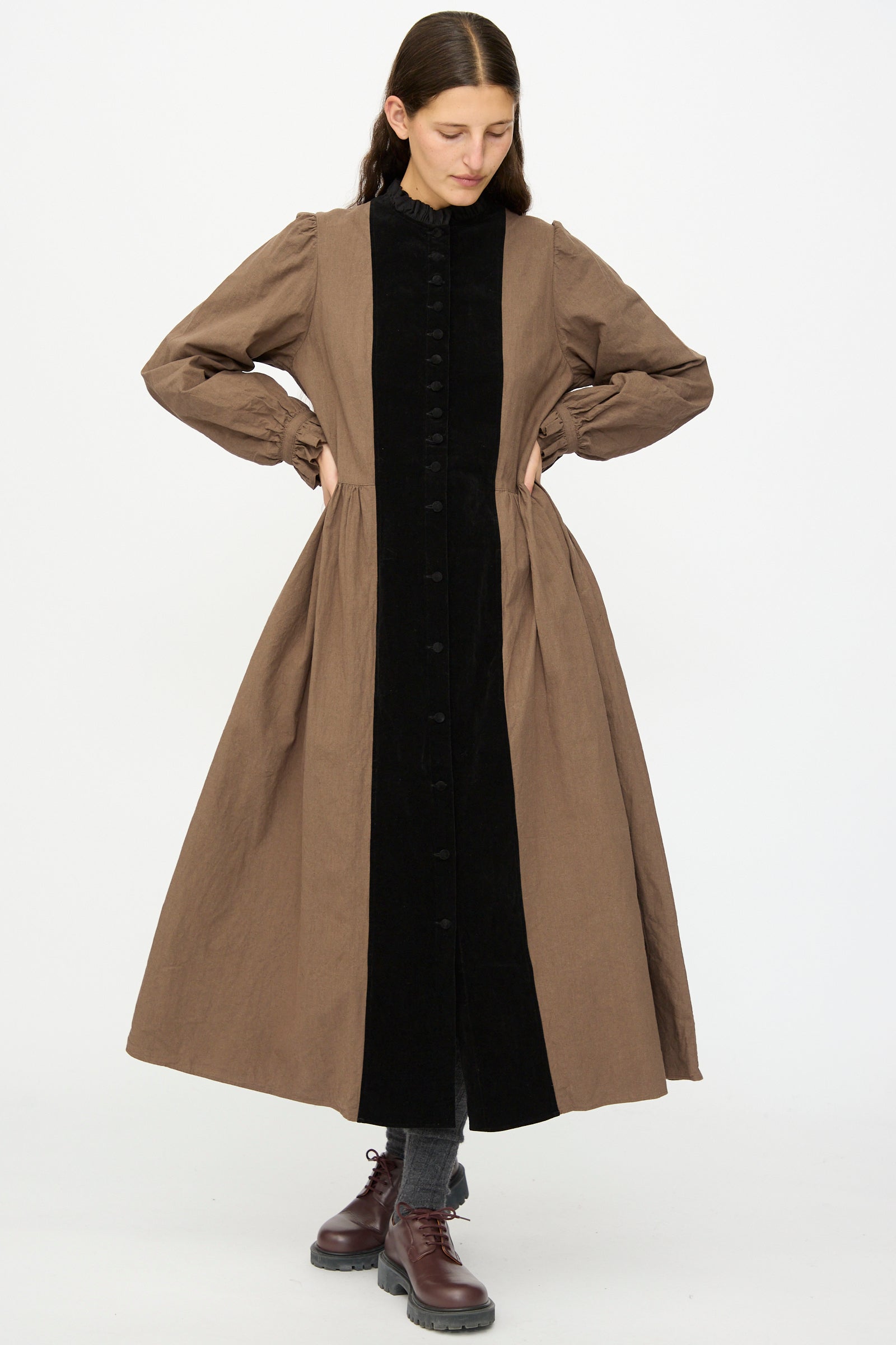 A person stands against a white background wearing Hallelujah’s Jeune Paysanne in Chocolate and Black, a long cotton dress made in Japan, paired with gray socks and brown lace-up shoes—an ensemble of understated elegance.