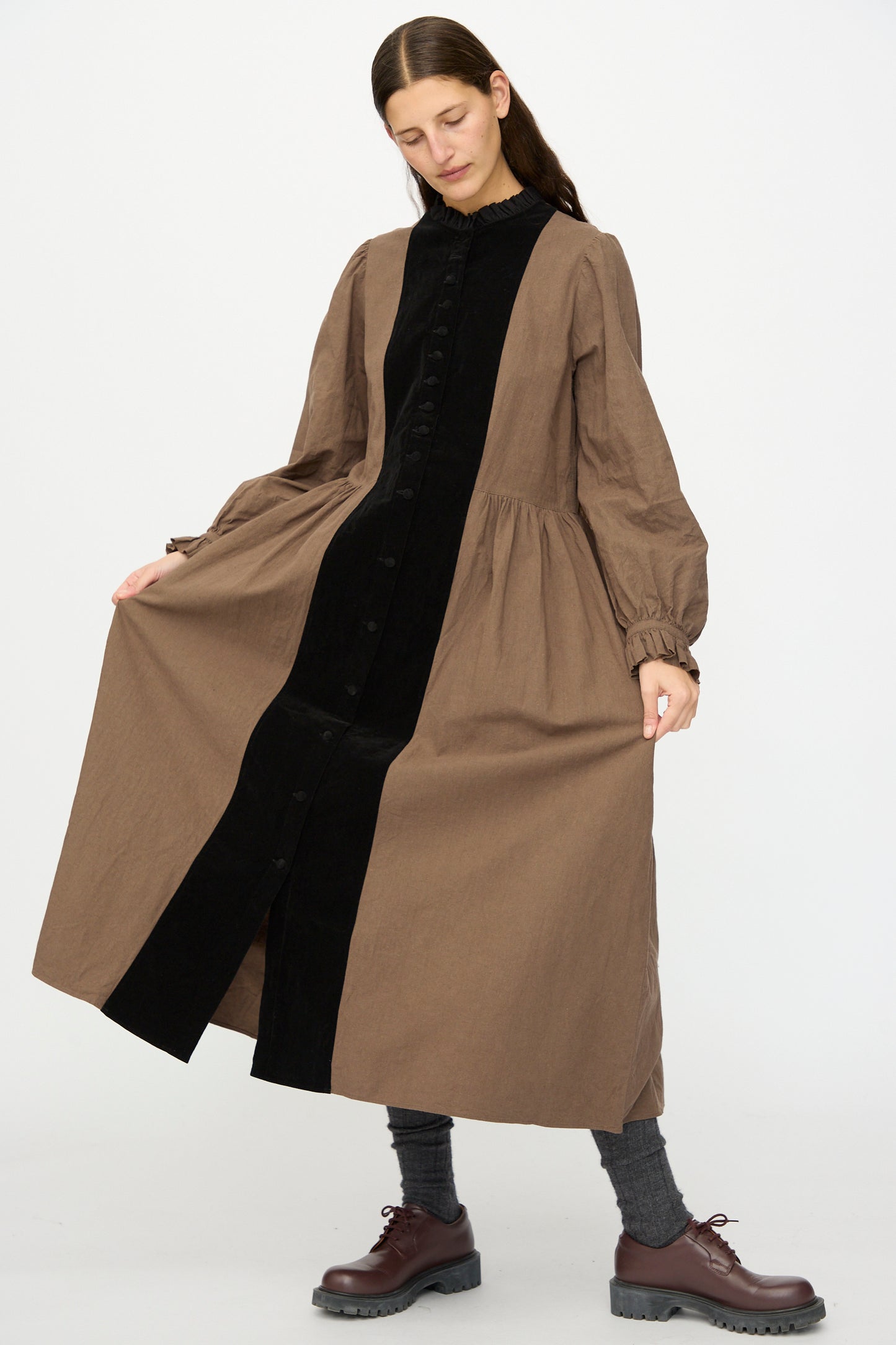 A person models the Jeune Paysanne in Chocolate and Black by Hallelujah, a stylish, Japanese-made cotton velveteen dress with black stripes and a ruffle collar, paired with gray socks and brown shoes against a white background.