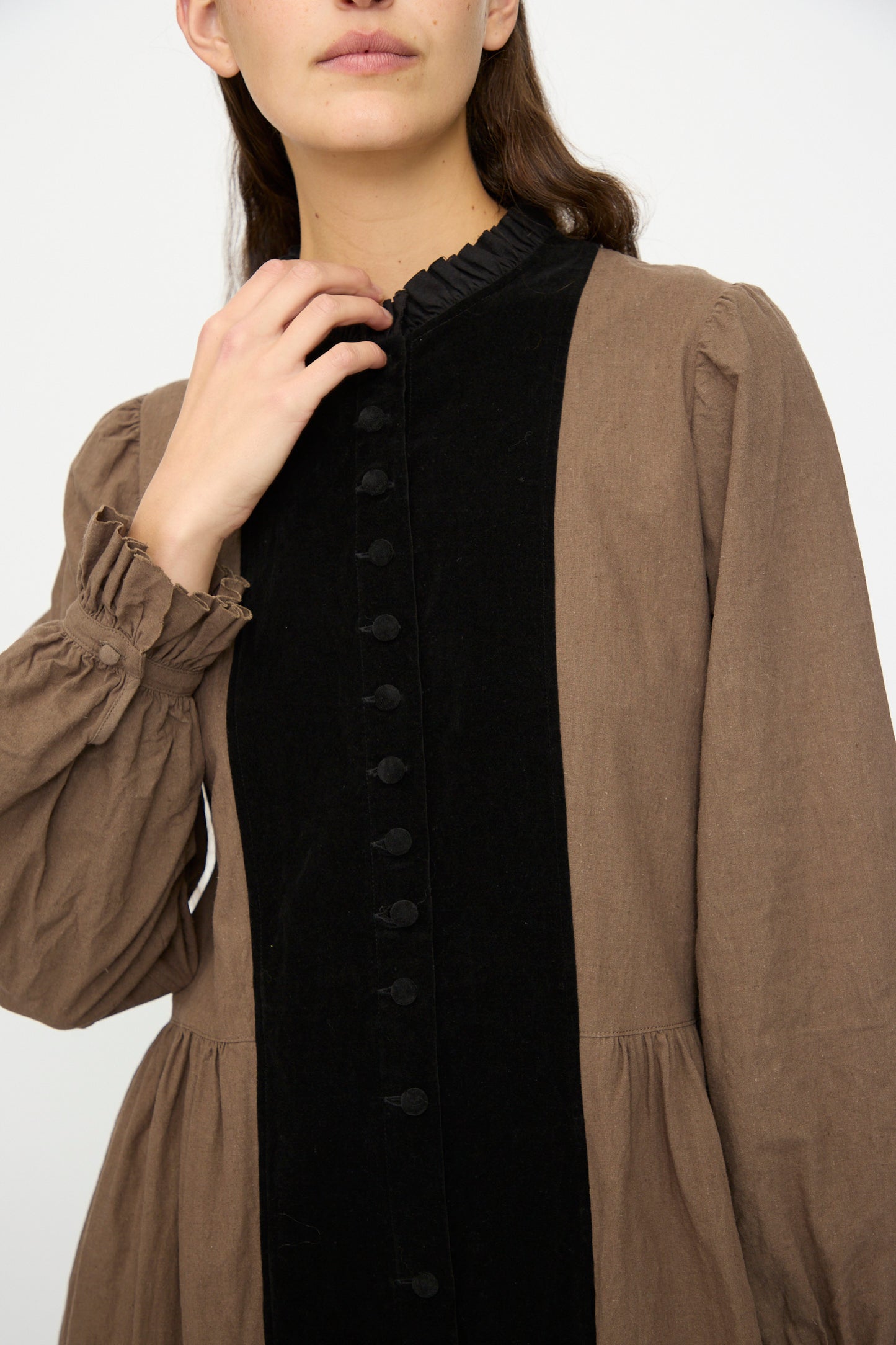 A person wearing Hallelujah’s Jeune Paysanne in Chocolate and Black, a long-sleeved velveteen cotton dress with a brown body and black buttoned panel, touches the collar with one hand.