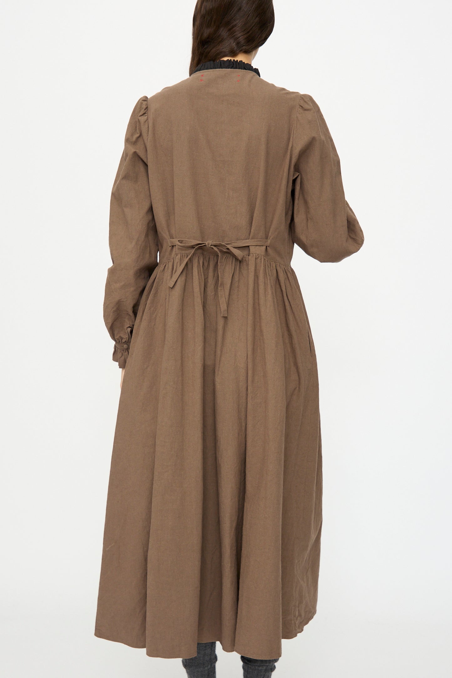 A person seen from the back wears the Hallelujah Jeune Paysanne dress in chocolate and black, featuring long sleeves, velveteen cotton, and a waist tie, set against a plain white background.