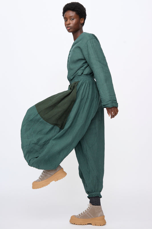 A model wears a green Linen Bloomer jumpsuit with a balloon silhouette, long sleeves, and tan lug-sole sneakers.