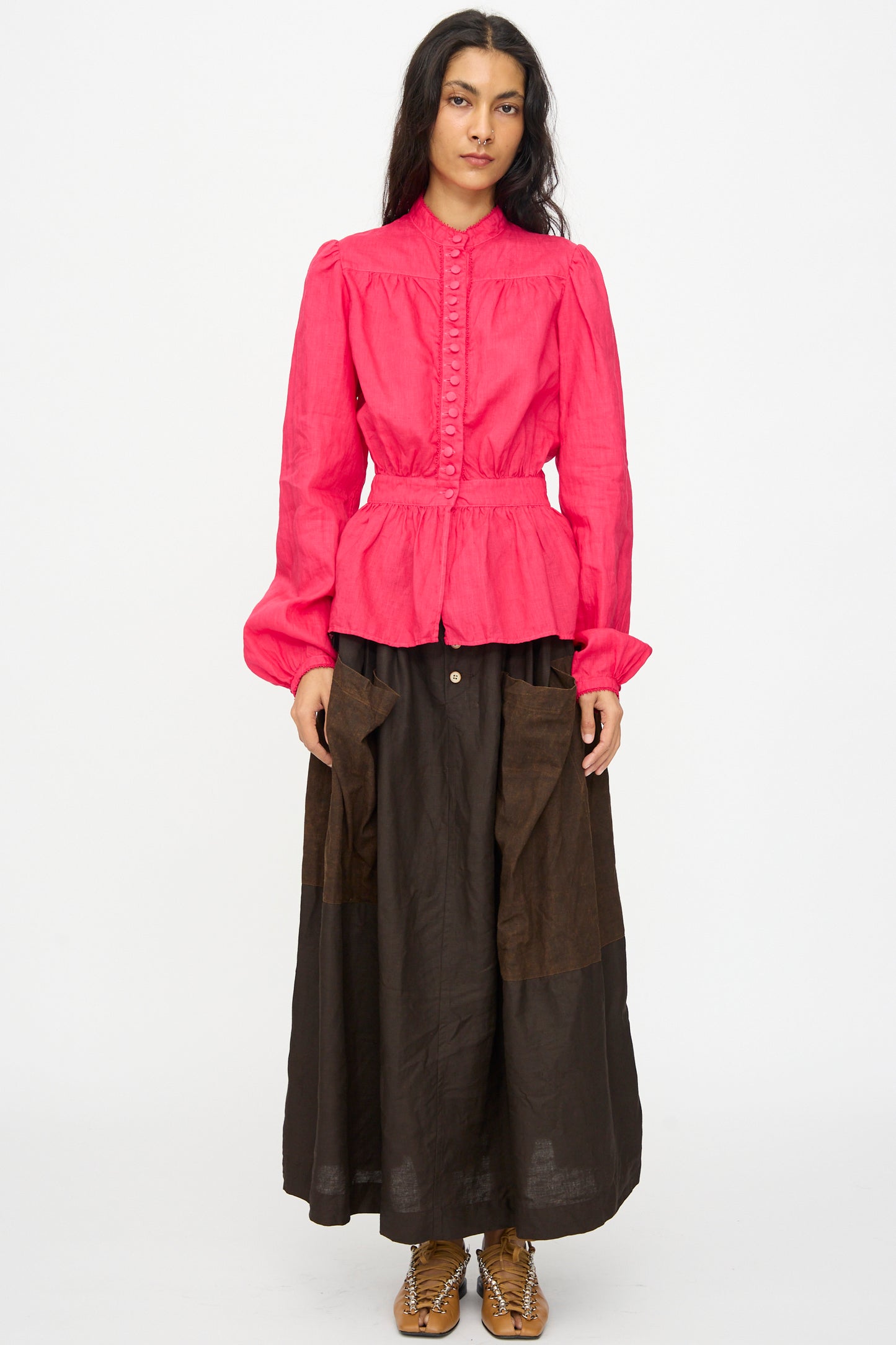 A person stands facing forward against a plain white background, wearing the Hallelujah Linen Jupe Bergère skirt in Iron Oxide, Ink and Persimmon Dye and a bright pink long-sleeve blouse with brown shoes.