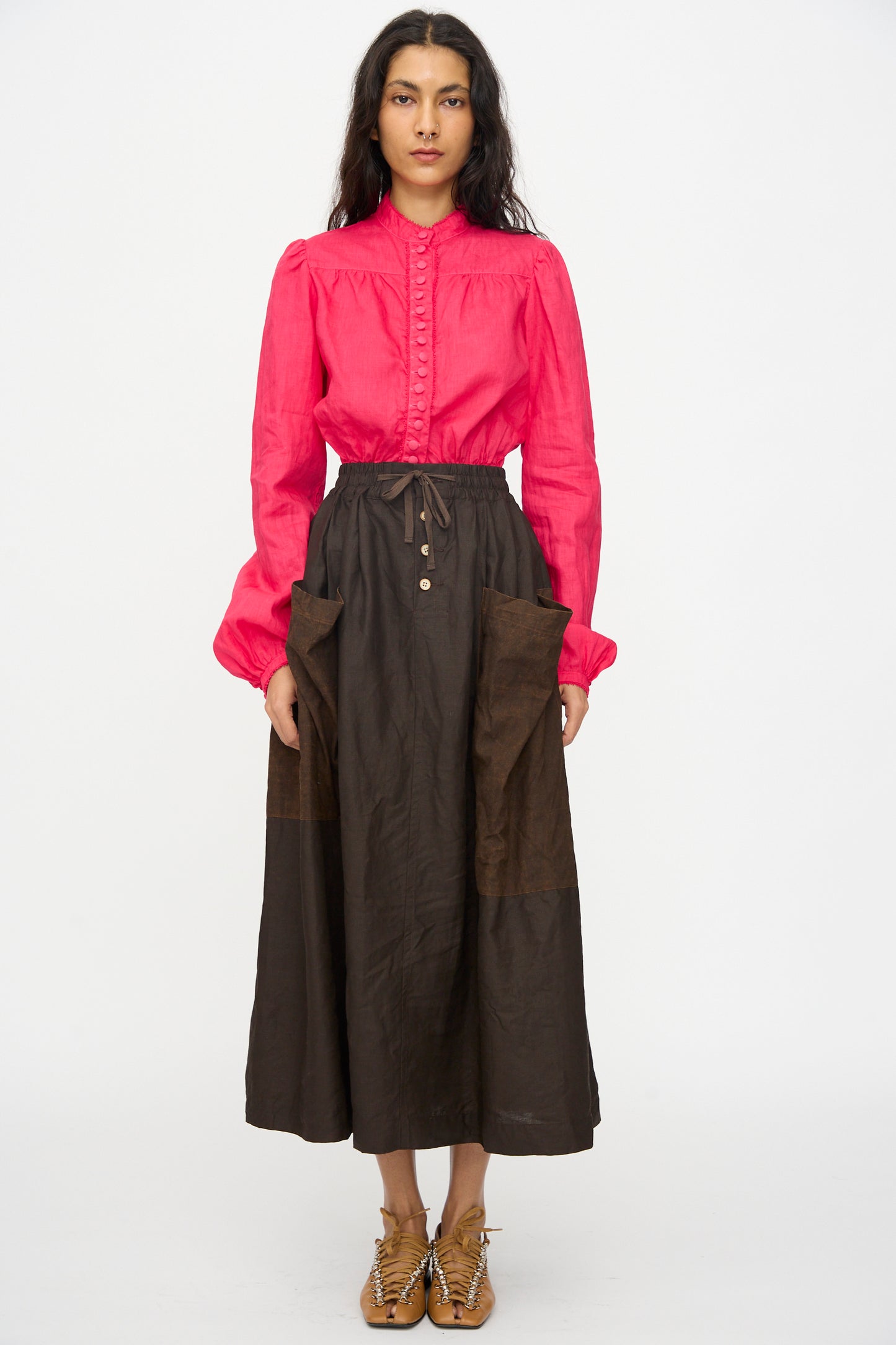 A woman stands facing forward, wearing a bright pink long-sleeve blouse and the Hallelujah Linen Jupe Bergère in Iron Oxide, Ink and Persimmon Dye—a dark brown A-line linen skirt with large pockets—paired with tan lace-up shoes.