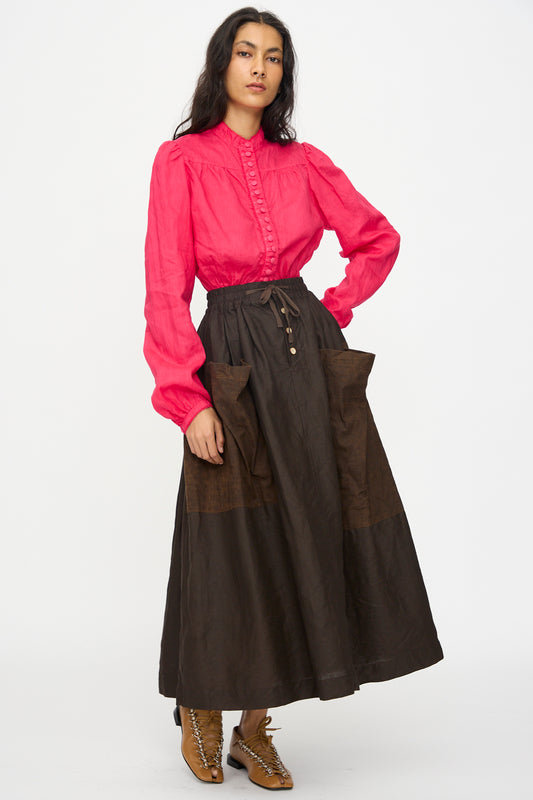A woman stands against a plain background wearing a bright pink long-sleeve blouse, tan lace-up boots, and the Hallelujah Linen Jupe Bergère high-waisted brown A-line skirt with large pockets in Iron Oxide, Ink and Persimmon Dye.