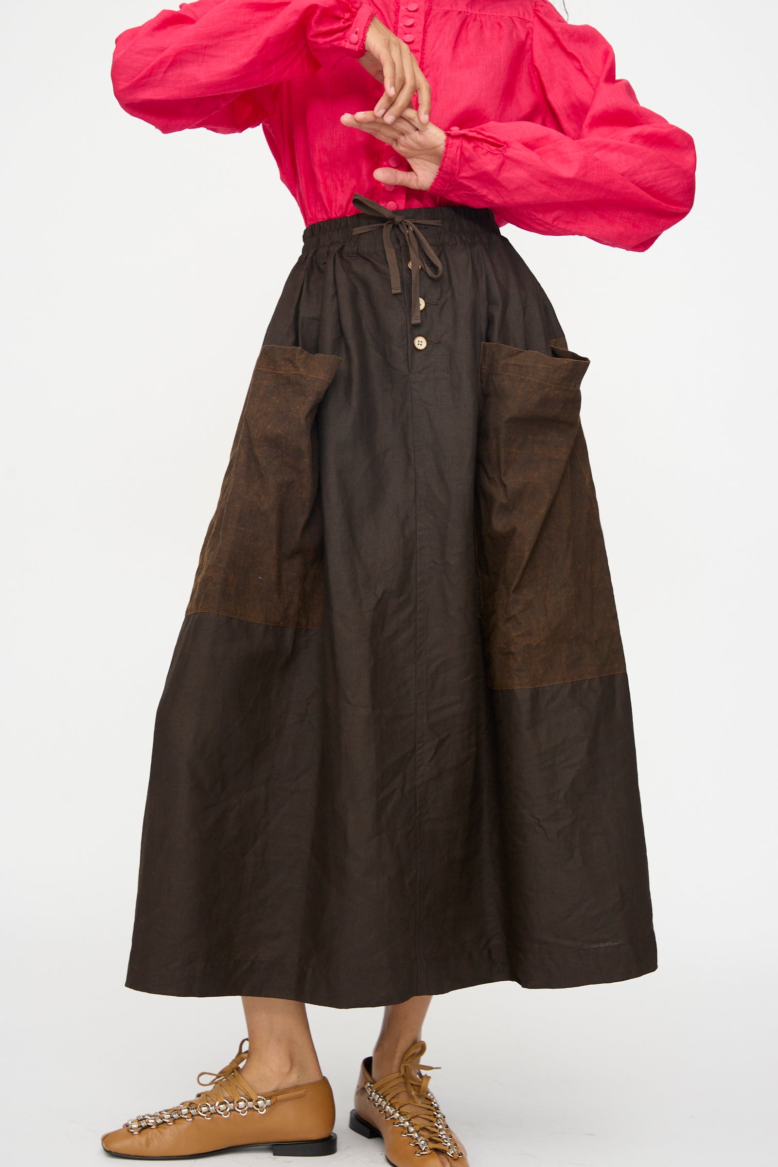 Model wearing Hallelujah's Linen Jupe Bergère skirt in Iron Oxide, Ink and Persimmon Dye, paired with a bright pink long-sleeve top and tan chain-detailed flats, posing against a plain white background.