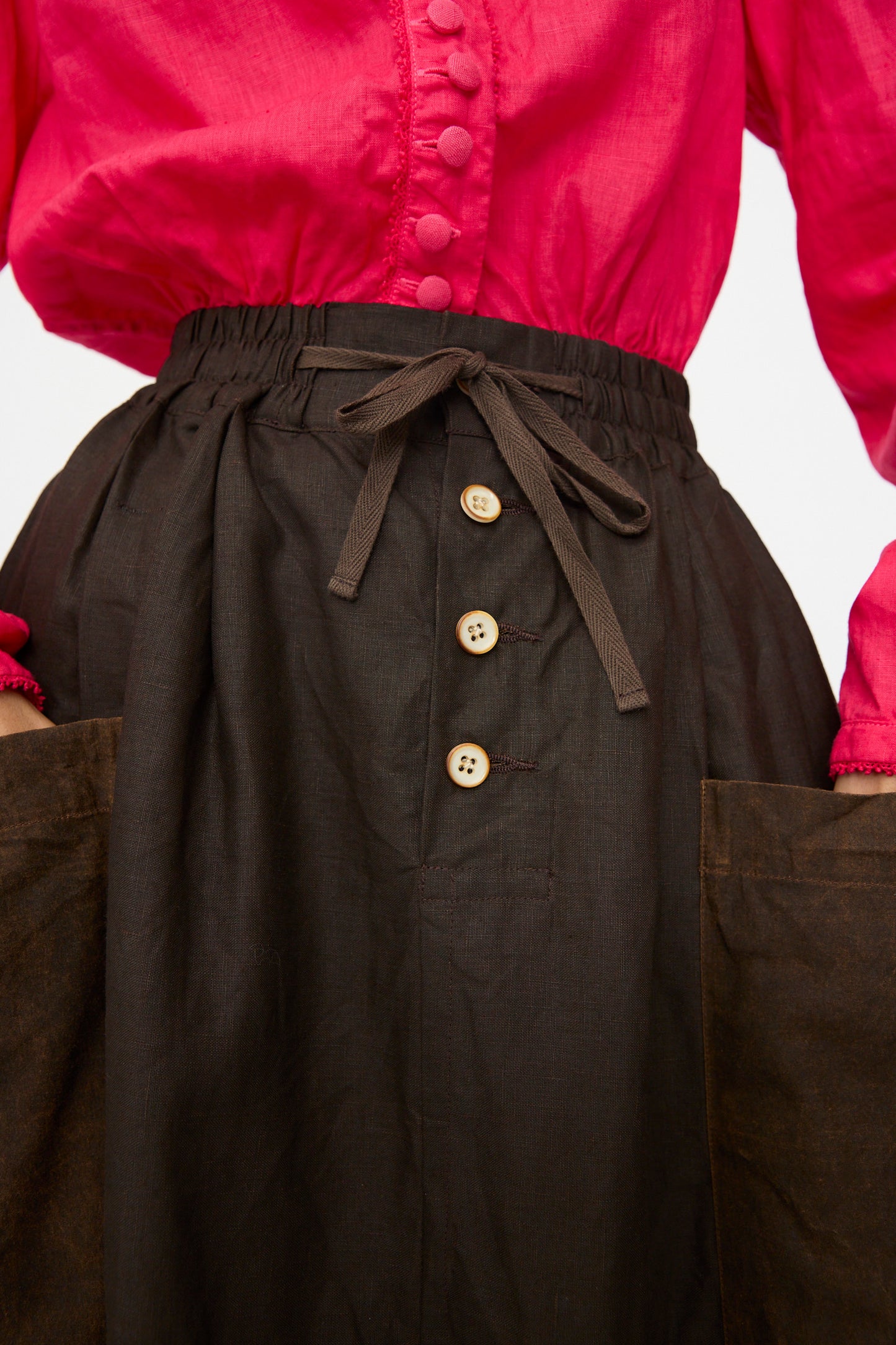 Person wearing a bright pink button-up shirt tucked into the Hallelujah Linen Jupe Bergère skirt in Iron Oxide, Ink and Persimmon Dye, which features a high waist, drawstring waist, button details, and large front pockets.