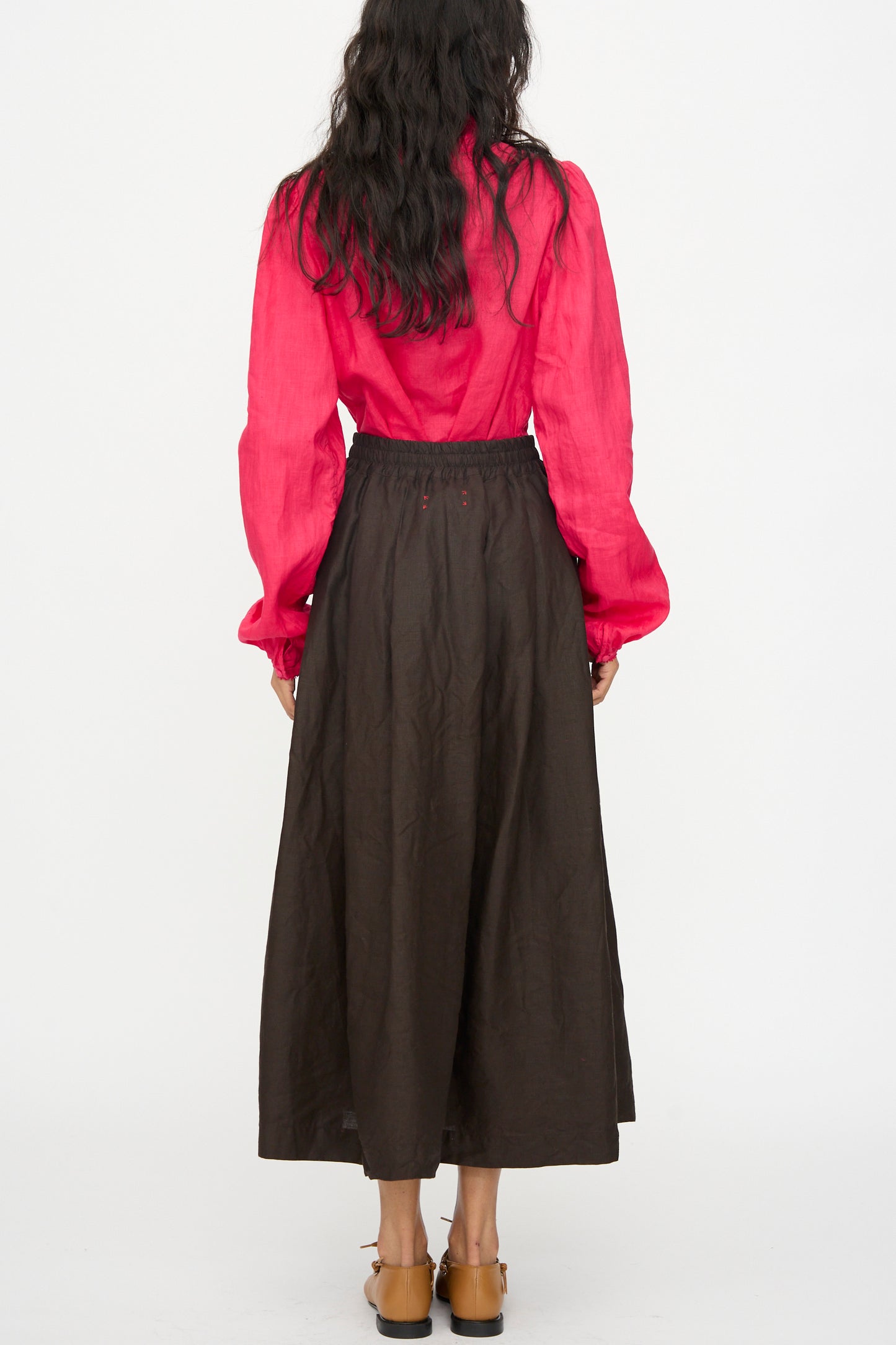 A person stands facing away, wearing a long-sleeve red blouse and Hallelujah's Linen Jupe Bergère skirt in Iron Oxide, Ink and Persimmon Dye, with tan shoes, set against a plain white background.