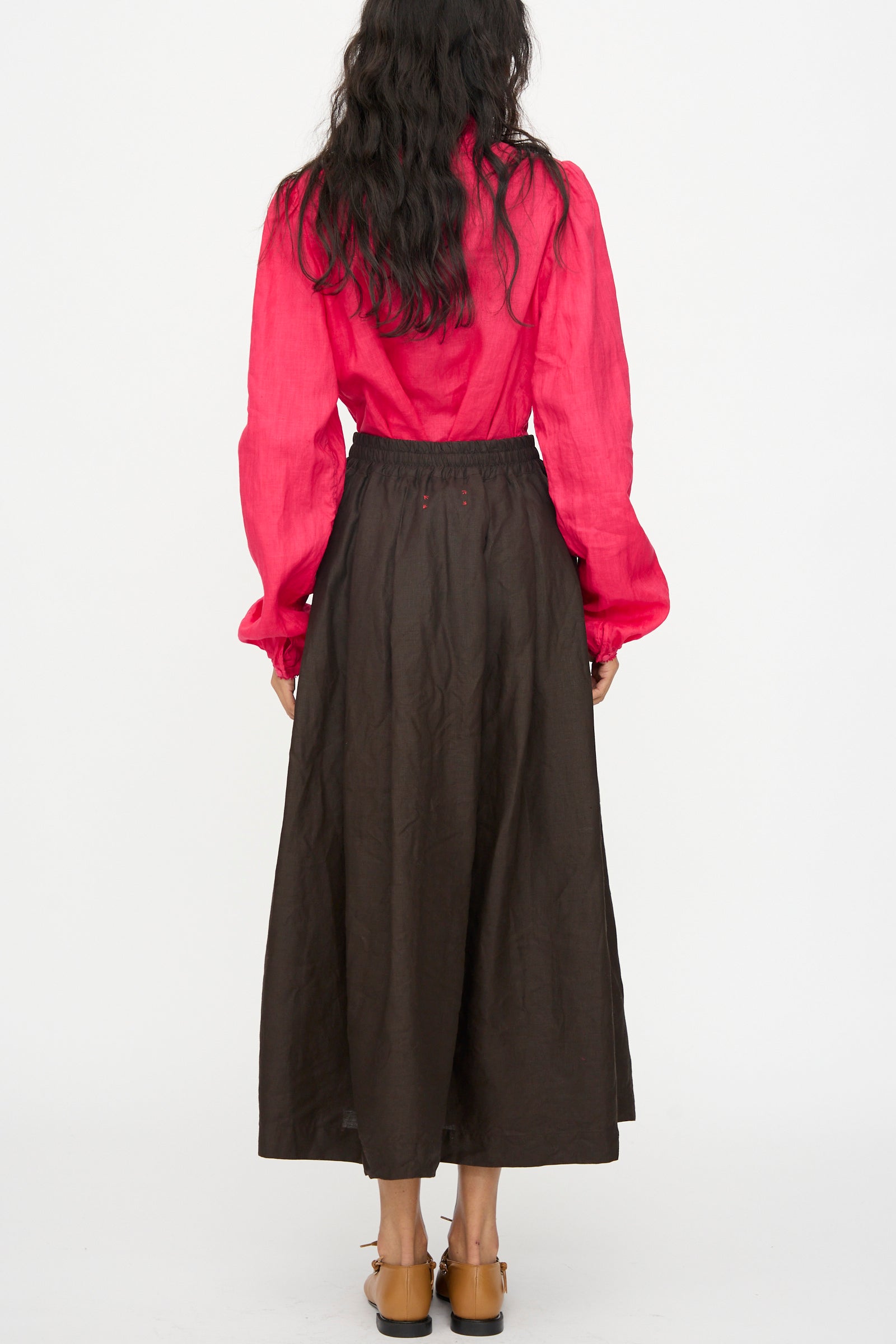 A person stands facing away, wearing a long-sleeve red blouse and Hallelujah's Linen Jupe Bergère skirt in Iron Oxide, Ink and Persimmon Dye, with tan shoes, set against a plain white background.