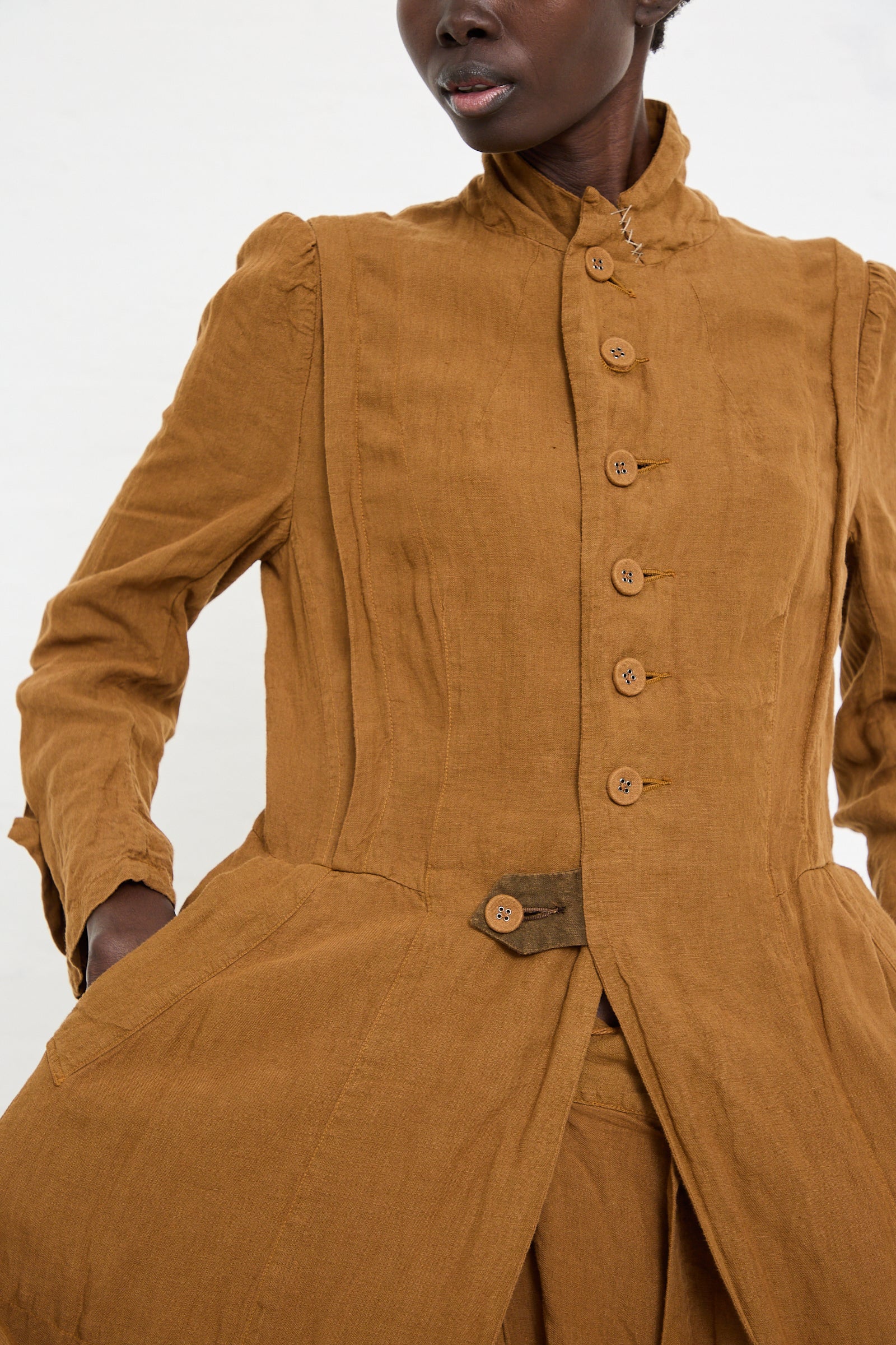 A person wearing a brown, high-collared button-up jacket with puffed shoulders and a flared waist, crafted using a unique dyeing process, the Linen Manteau de Pompier 1800's in Coffee by Hallelujah.