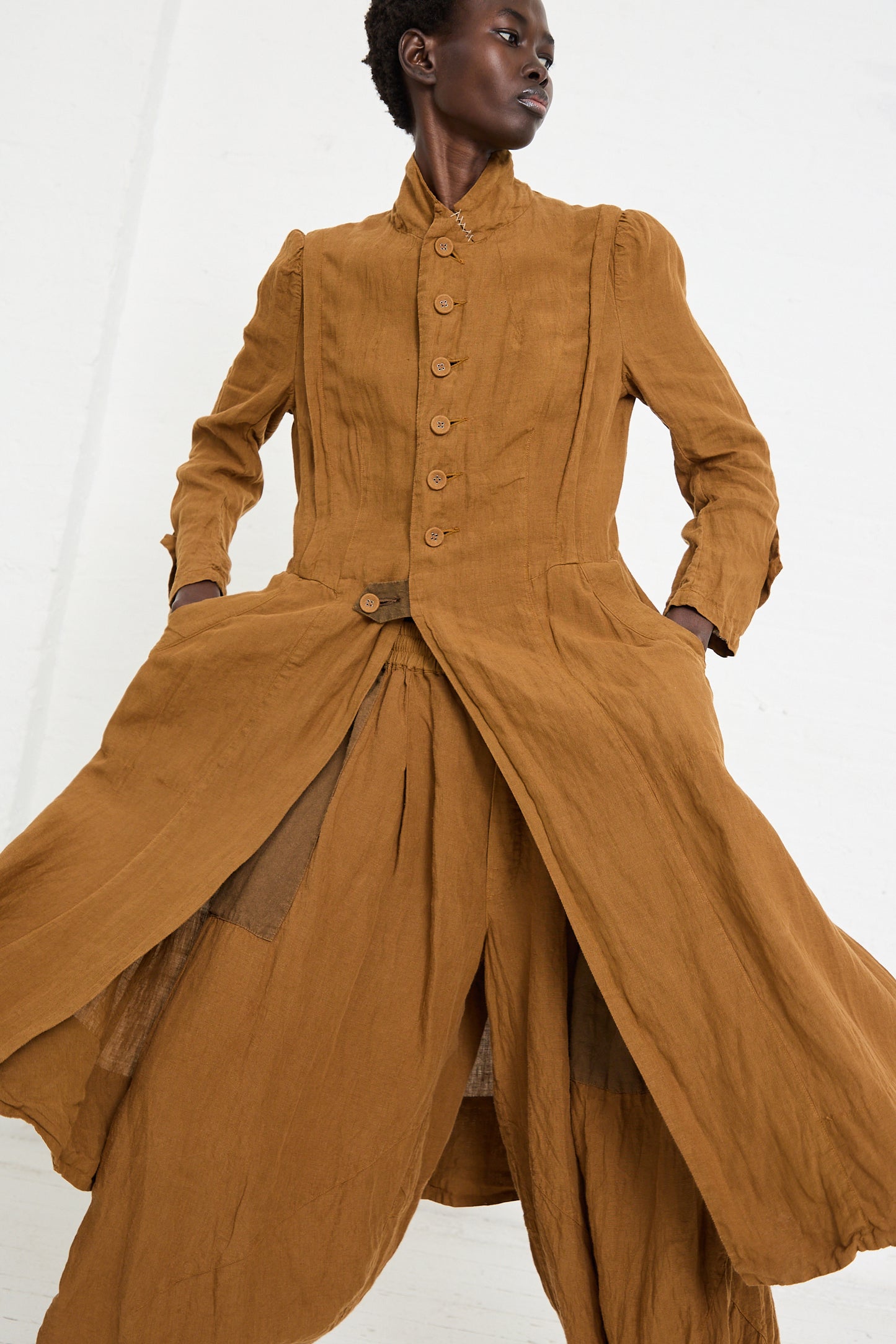 A person stands with hands in pockets, wearing the Hallelujah Linen Manteau de Pompier 1800's in Coffee. The long brown dress boasts layered fabric and a high collar. The background is plain white.