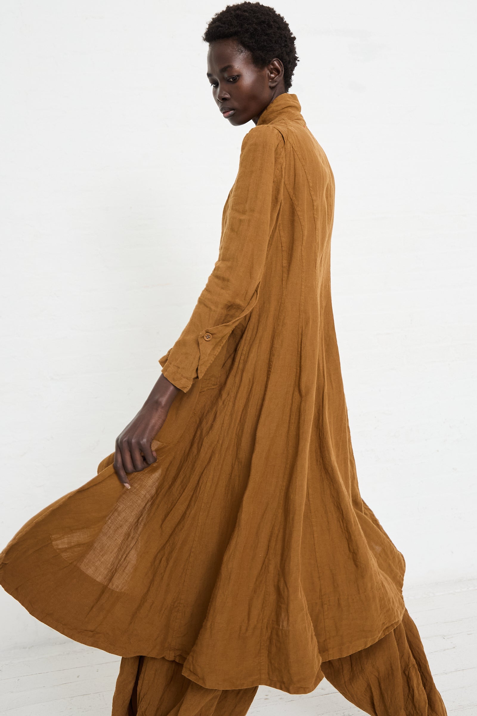 A person with short hair wears a long, flowing brown dress and matching pants, each piece handmade in Kyoto. They walk away from the camera in front of a plain white background, wearing the Linen Manteau de Pompier 1800's in Coffee by Hallelujah.