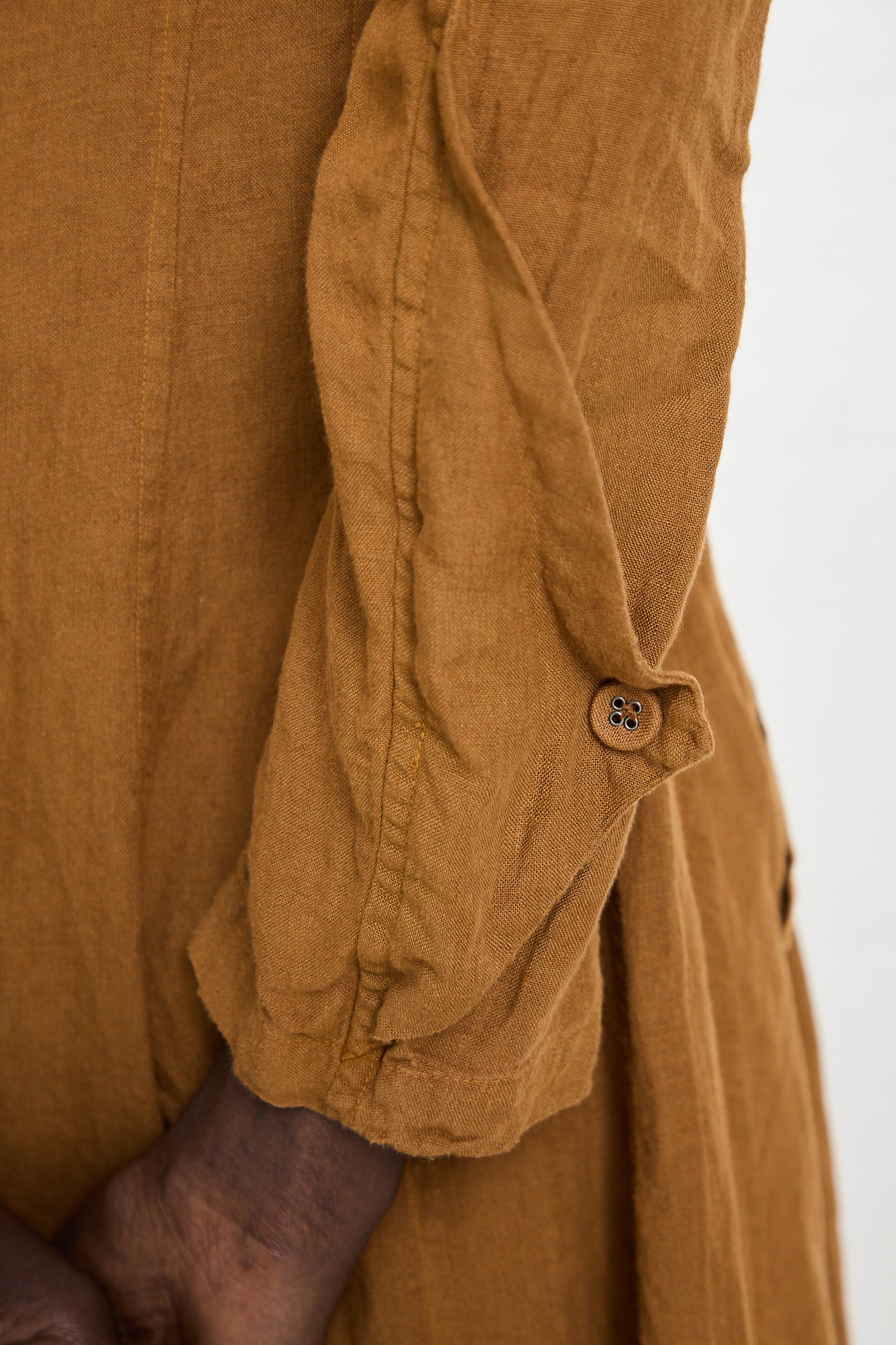 Close-up of a person wearing a long-sleeved, wrinkled, light brown Hallelujah Linen Manteau de Pompier 1800's in Coffee handmade in Kyoto. The sleeve is partially rolled up and secured with a buttoned tab. The person's hands are crossed at the waist.