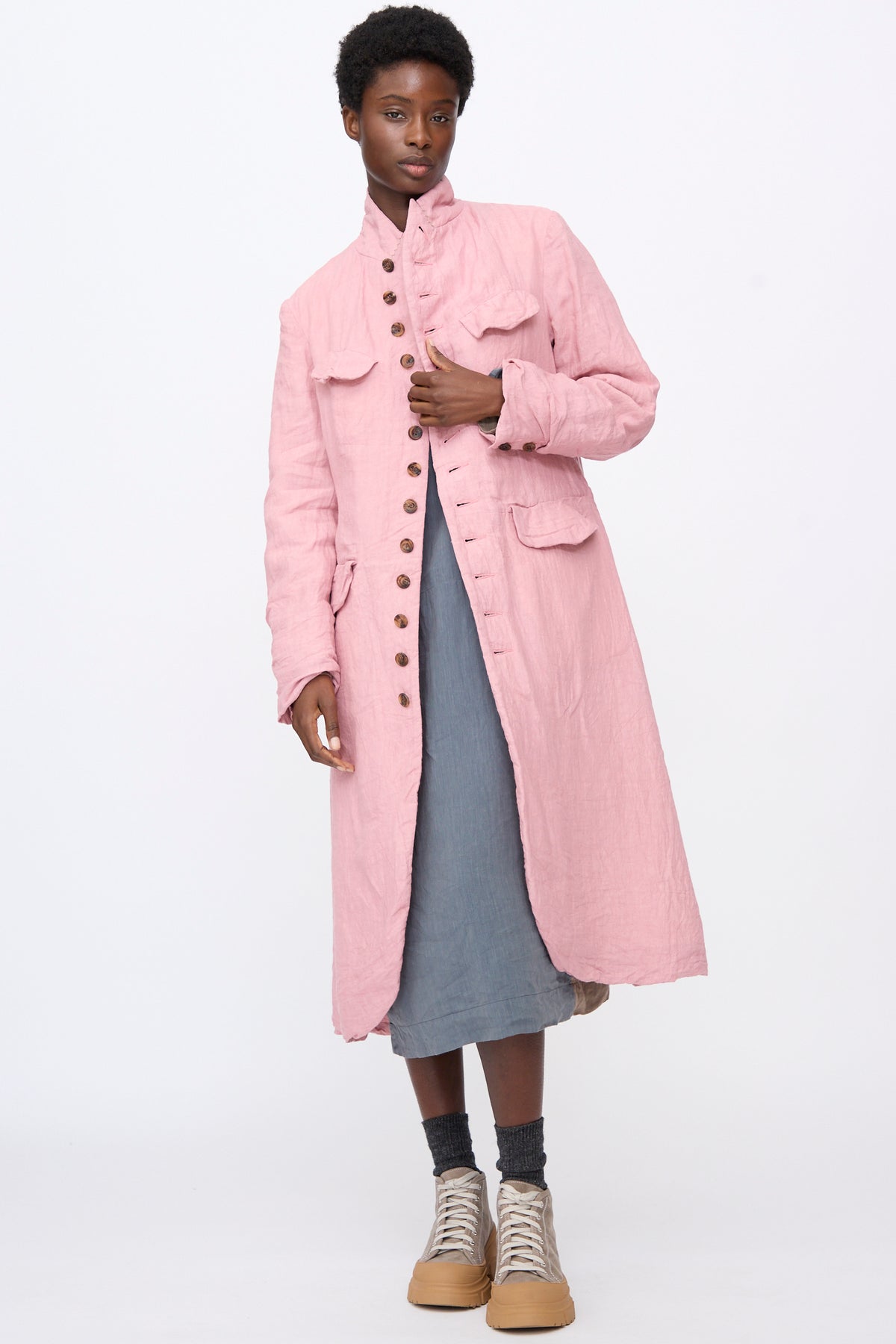 A person wearing a relaxed fit, pink Linen Manteaux de Chasse over a blue dress, with grey socks and beige sneakers.