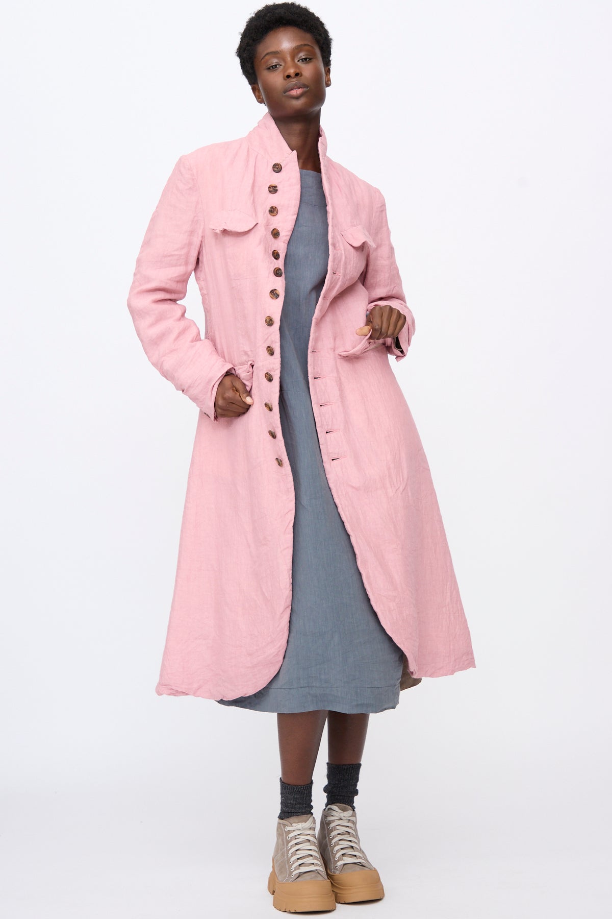 A person wearing a relaxed fit, pink Linen Manteaux de Chasse over a grey dress and sneakers.