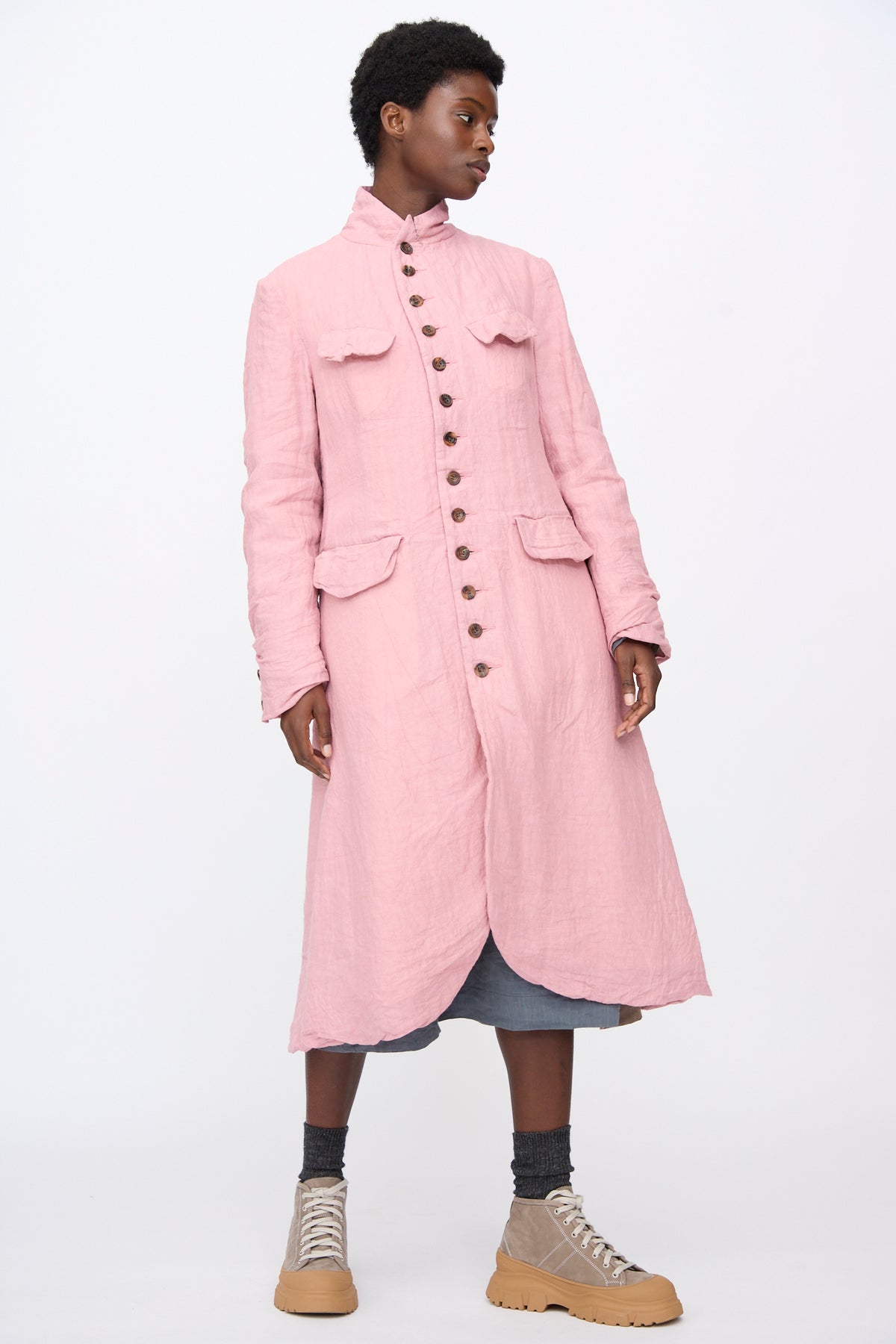 A person wears a long, pink, relaxed-fit Hallelujah Linen Manteaux de Chasse linen hunting coat with flap pockets.