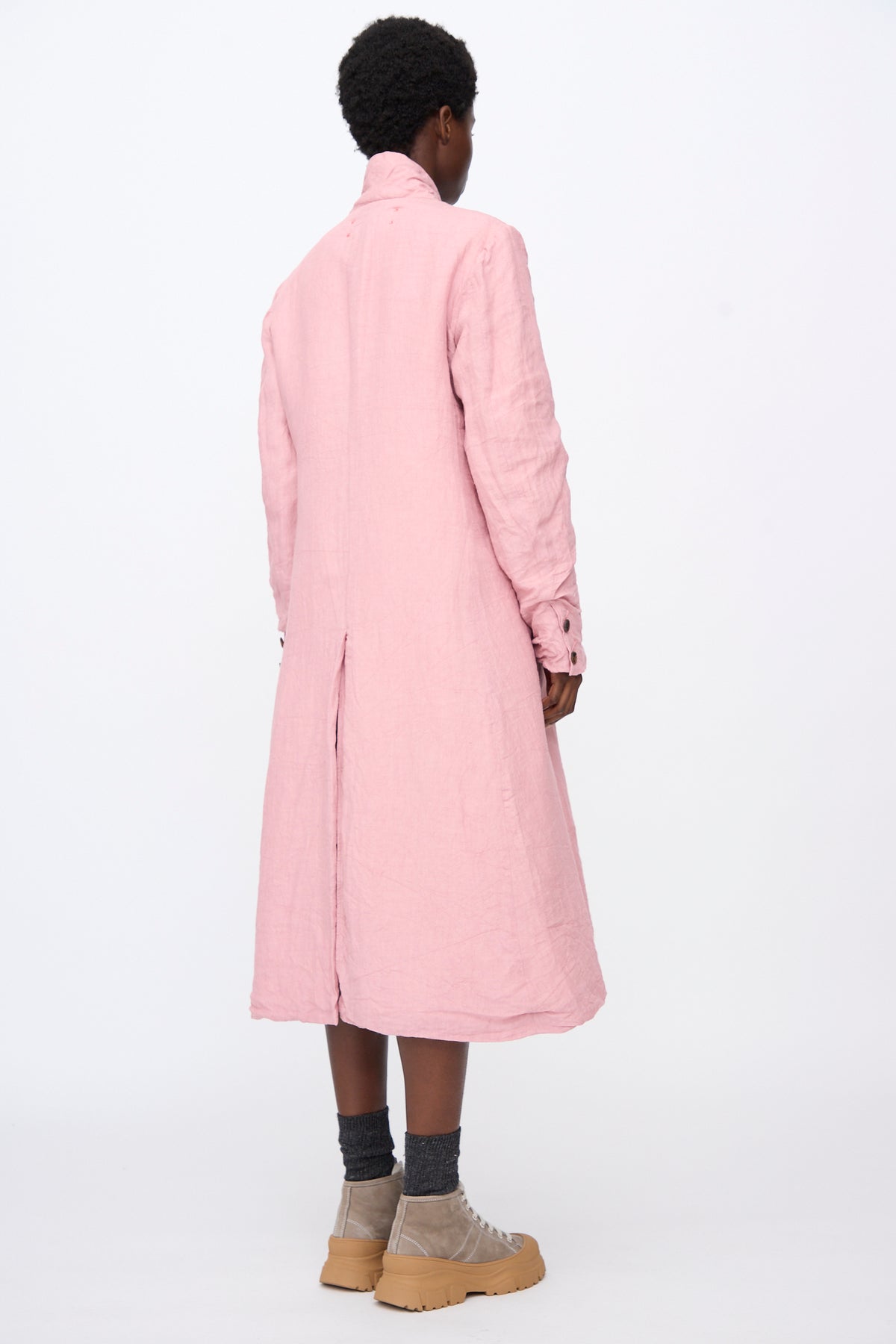 A person seen from behind wears a relaxed fit pink Hallelujah Linen Manteaux de Chasse with a back vent, grey socks, and tan shoes with thick soles.