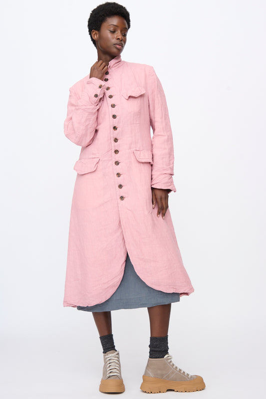 A person wears a relaxed fit pink Hallelujah Linen Manteaux de Chasse button-down coat over a grey skirt, dark socks, and tan lace-up sneakers.