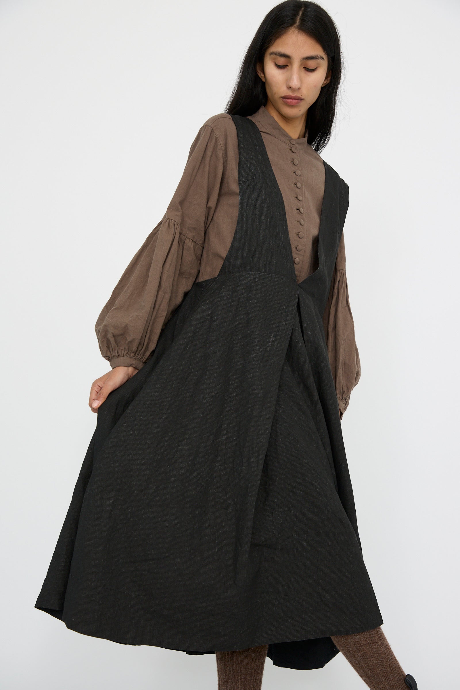 A person wearing Hallelujah's Linen Priest's Tablier in Charcoal Black over a brown blouse with puffy sleeves poses against a plain white background., looking down.