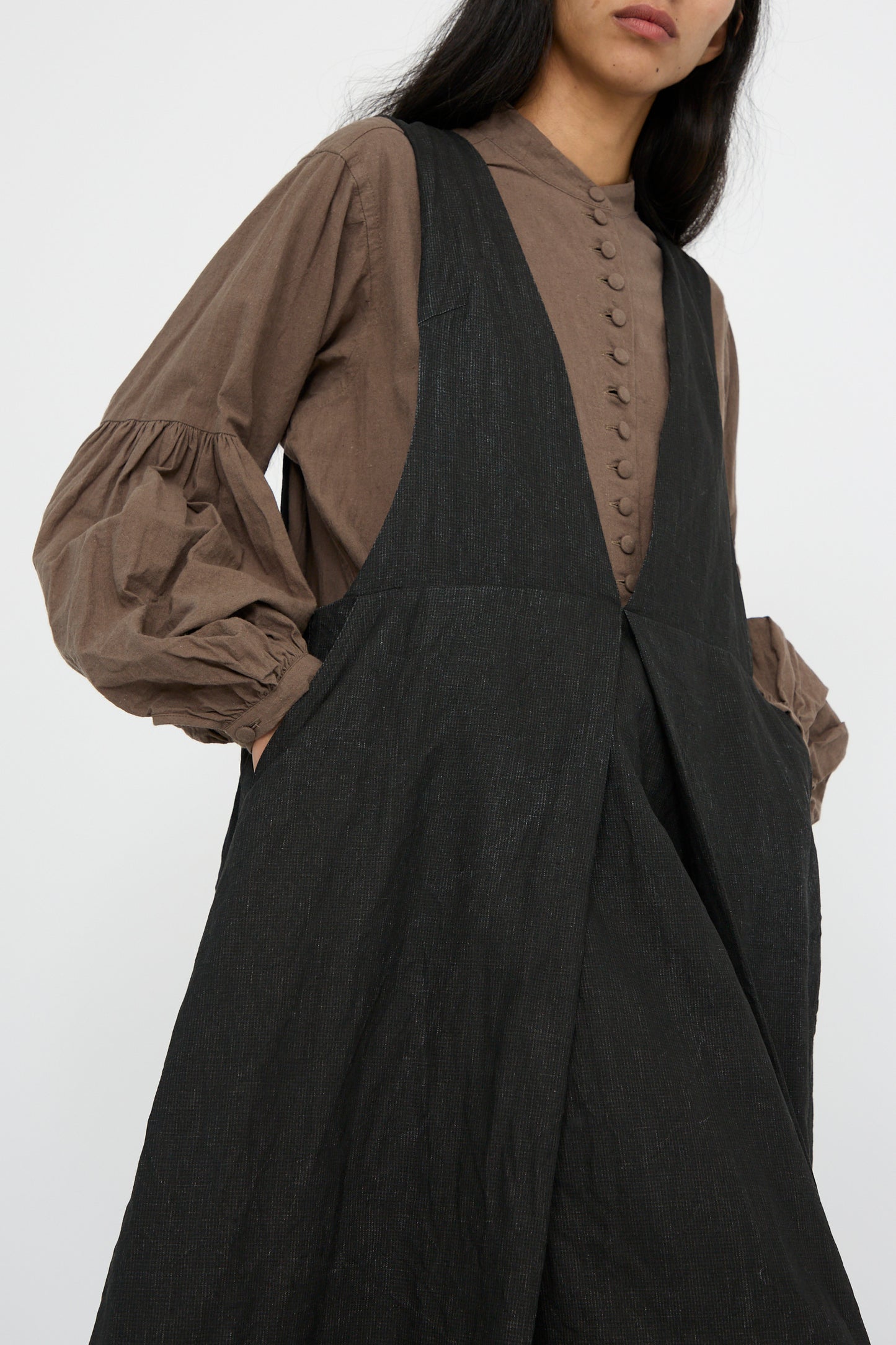 A person wearing a brown button-up blouse with puff sleeves under the Hallelujah Linen Priest's Tablier in charcoal black, hands in their pockets against a white background. 