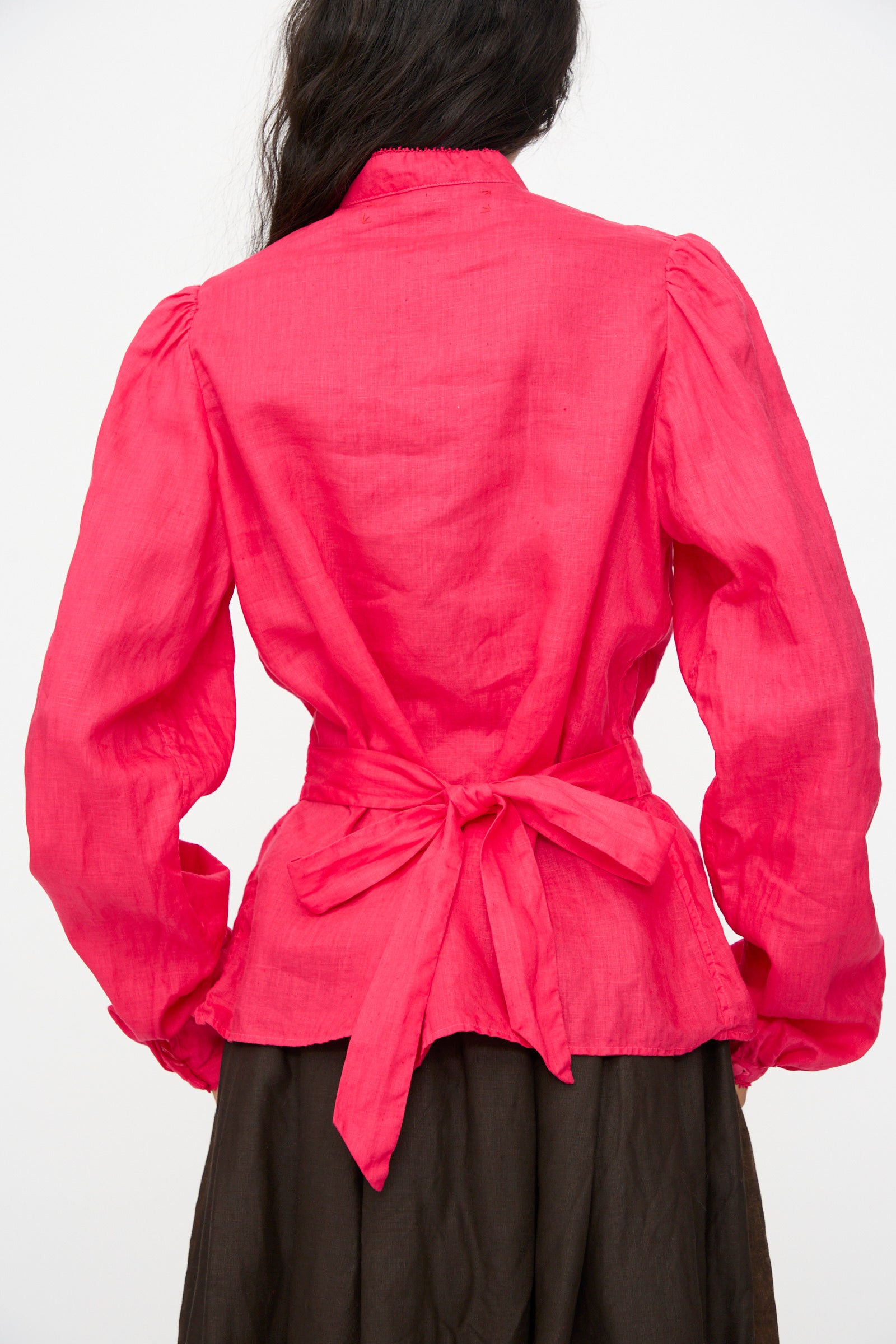A person is seen from the back wearing Hallelujah's Linen Réplique de Chemise Victorienne in Cherry Red, a long sleeve blouse with a tied waist bow, paired with a dark skirt. The stylish top is expertly made in Japan.