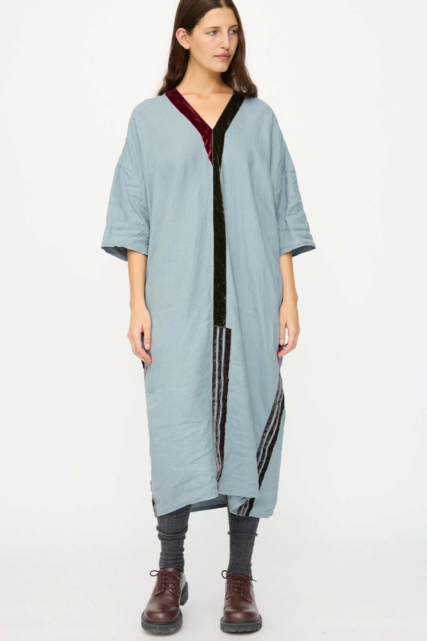 A woman stands against a plain white background wearing the Hallelujah Linen Robes de couture 1930s in Blue, featuring a relaxed fit and dark velvet trim, paired with gray leggings and brown lace-up shoes.