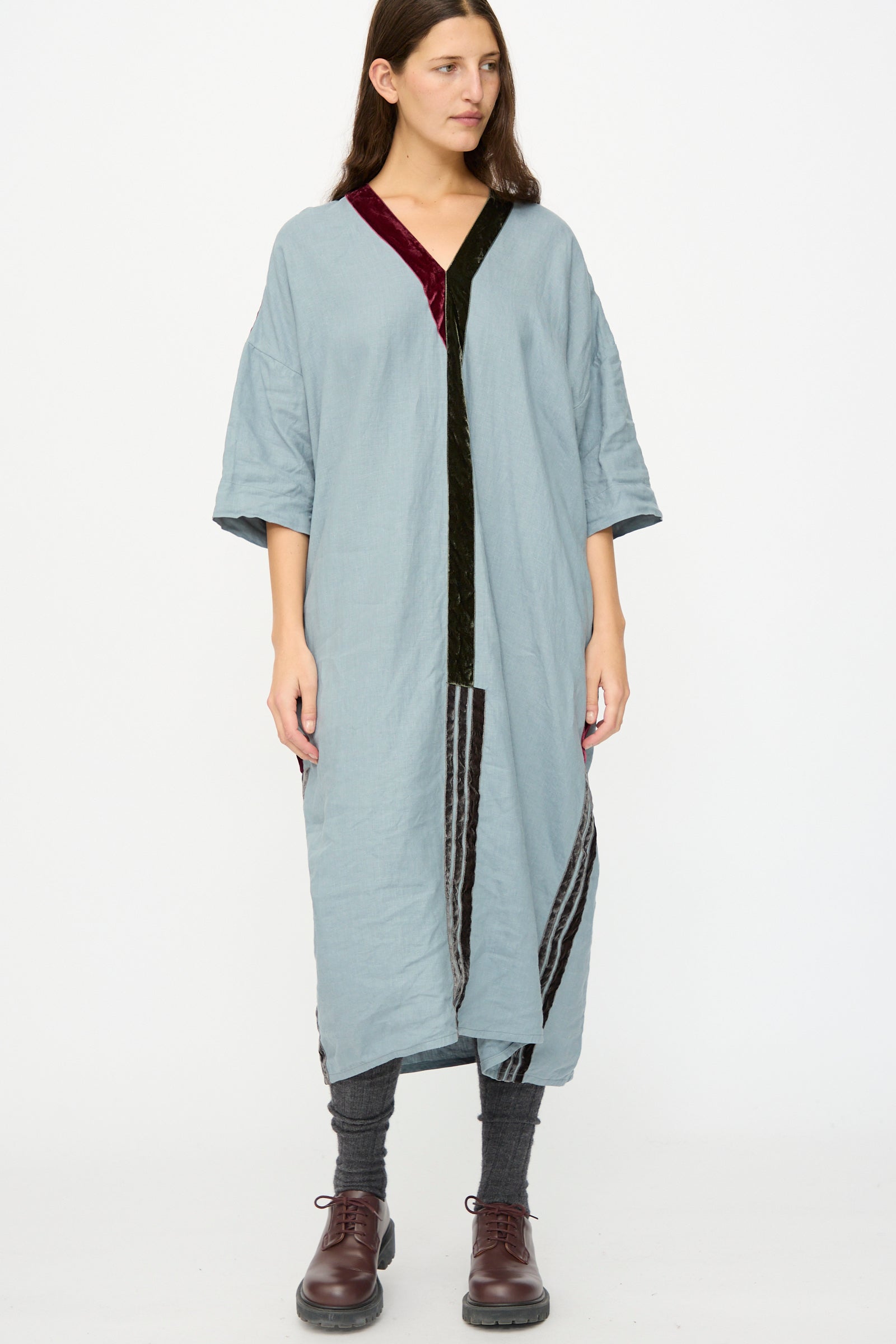 A woman stands against a plain white background wearing the Hallelujah Linen Robes de couture 1930s in Blue, featuring a relaxed fit and dark velvet trim, paired with gray leggings and brown lace-up shoes.
