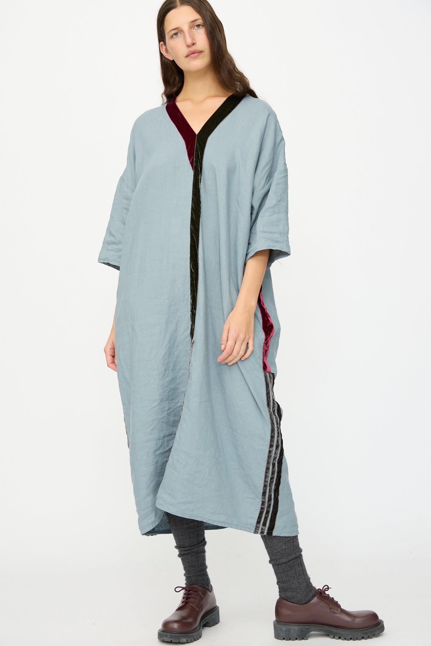 A person wears the Hallelujah Linen Robes de couture 1930s in Blue, featuring black and burgundy trim, paired with gray leggings and brown shoes, standing against a plain white background.