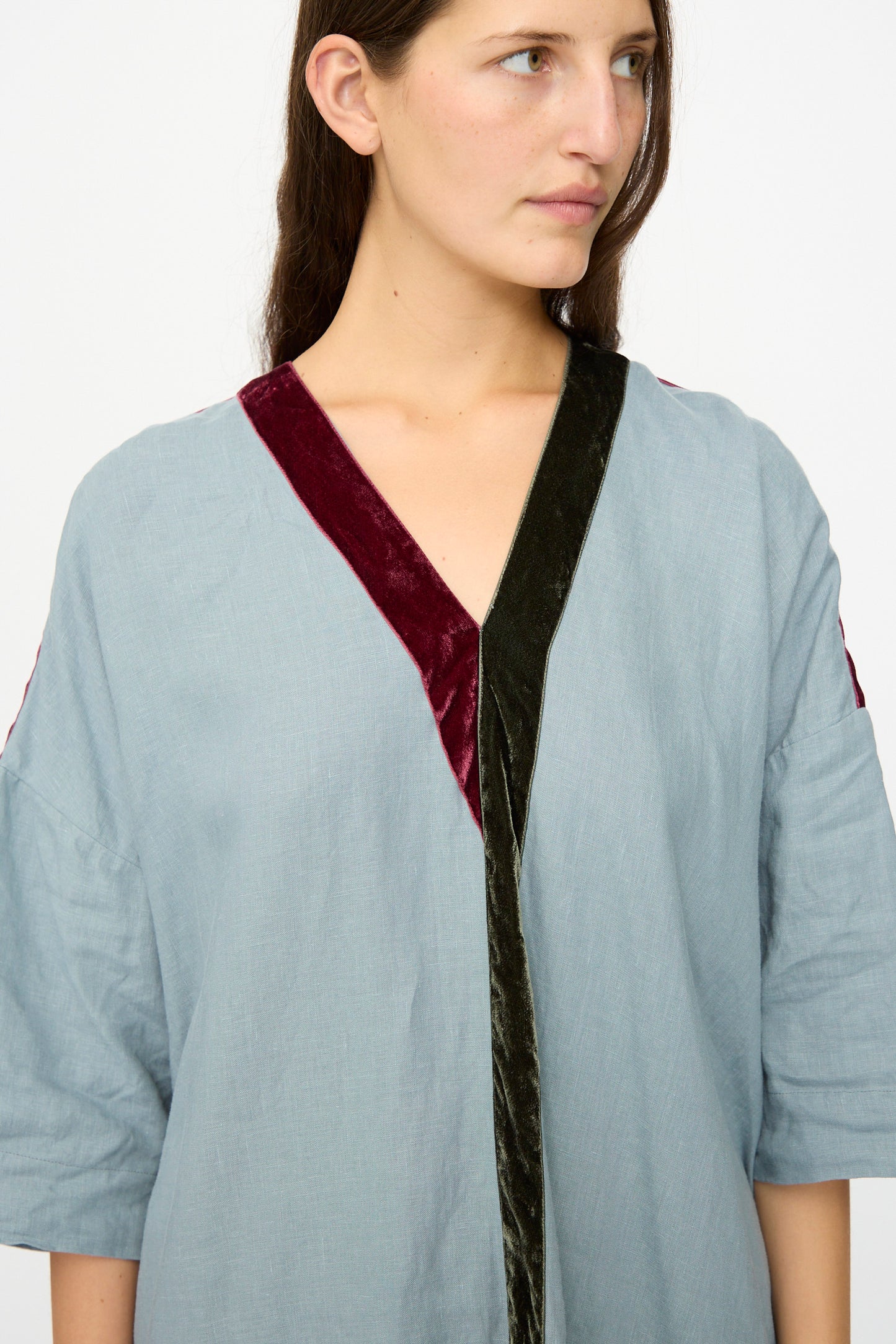 A woman in a Hallelujah Linen Robes de couture 1930s Blue dress, featuring a light blue V-neck and burgundy and dark green velvet trim, looks to the side against a plain background.