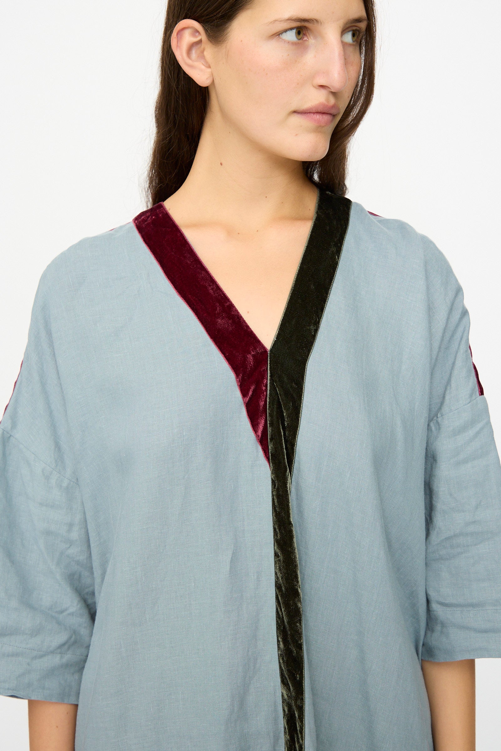 A woman in a Hallelujah Linen Robes de couture 1930s Blue dress, featuring a light blue V-neck and burgundy and dark green velvet trim, looks to the side against a plain background.