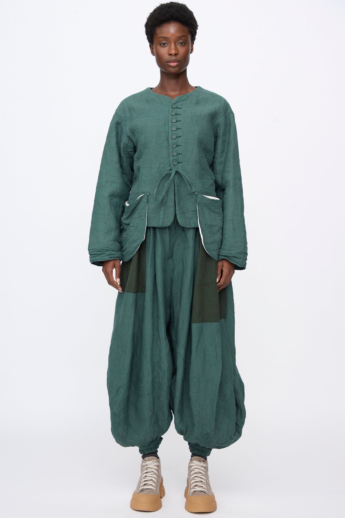 A person wearing a forest green Hallelujah Linen Veste de Berger jacket with a tie waist and matching wide-leg, cropped pants featuring contrast panel details.