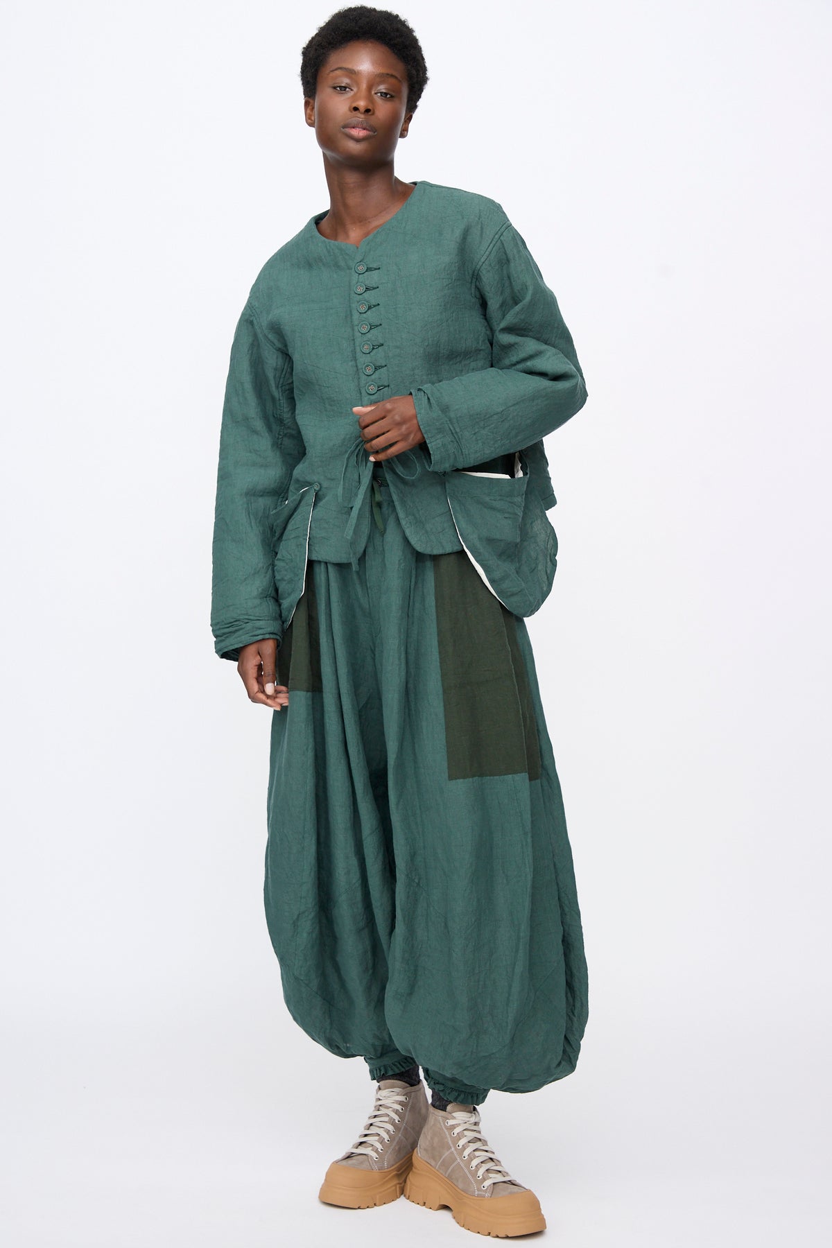 A person wearing a Hallelujah Linen Veste de Berger in Green, a collarless button-front jacket with matching loose-fitting, wide-leg trousers, styled with beige sneakers.