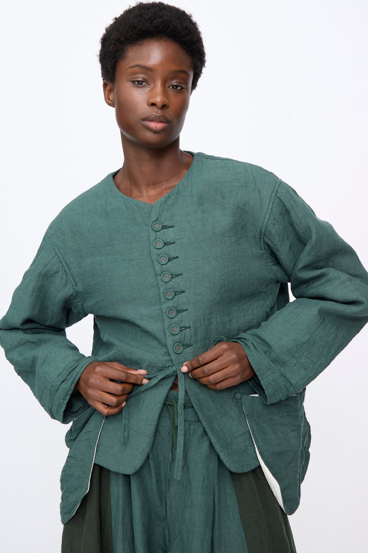 A model wearing a dark green Hallelujah Linen Veste de Berger with a high button-up collar and a waist tie.