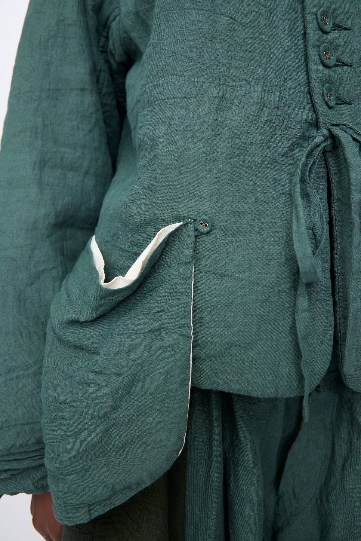 Hallelujah Linen Veste de Berger in Green, a jacket featuring front buttons, a waist tie, and a large side pocket with a light-colored lining.
