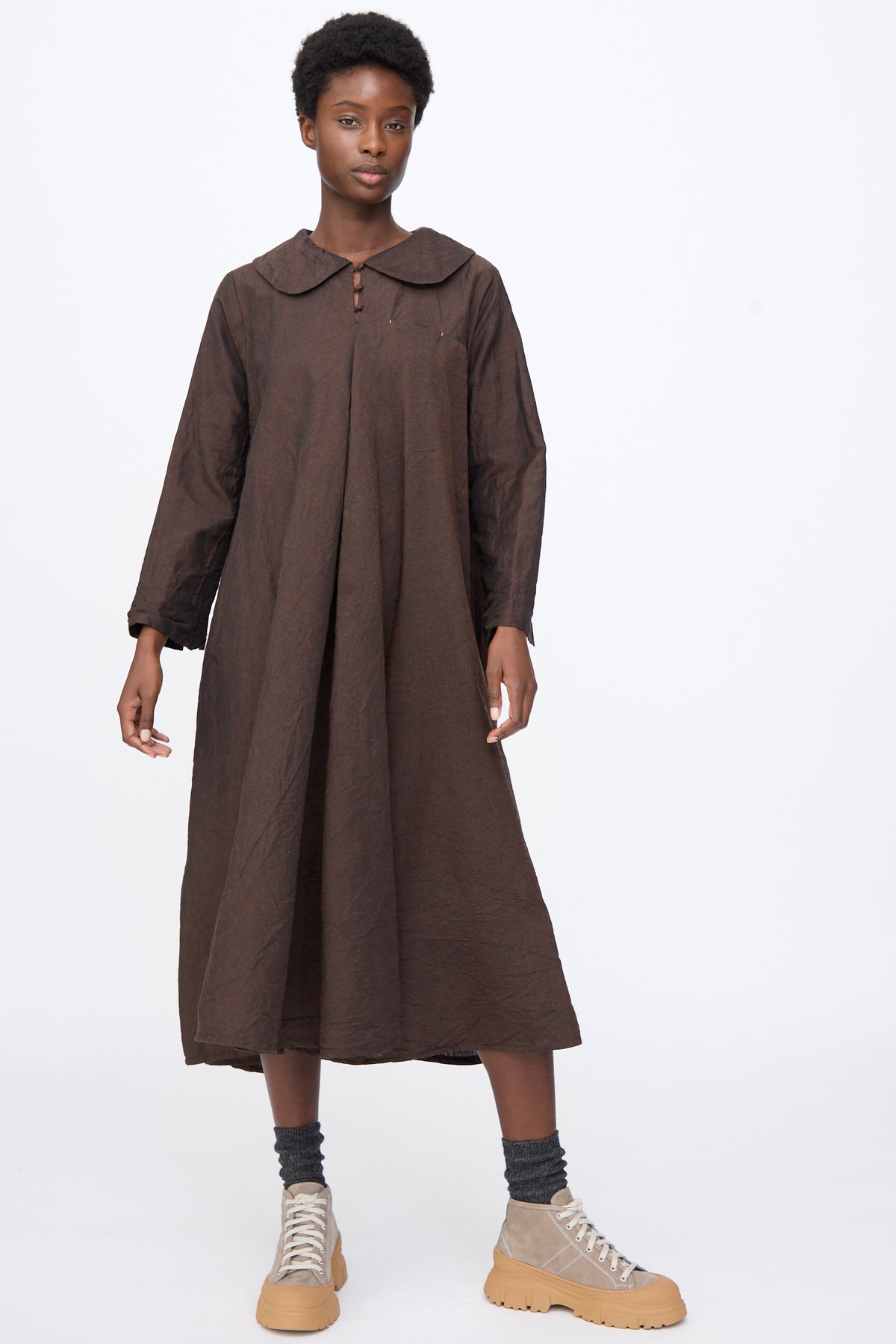 A person wears a Hallelujah Linen Wool Robe à Col Claudine in brown, a long-sleeve, calf-length dress with a rounded collar, grey socks, and tan platform sneakers.