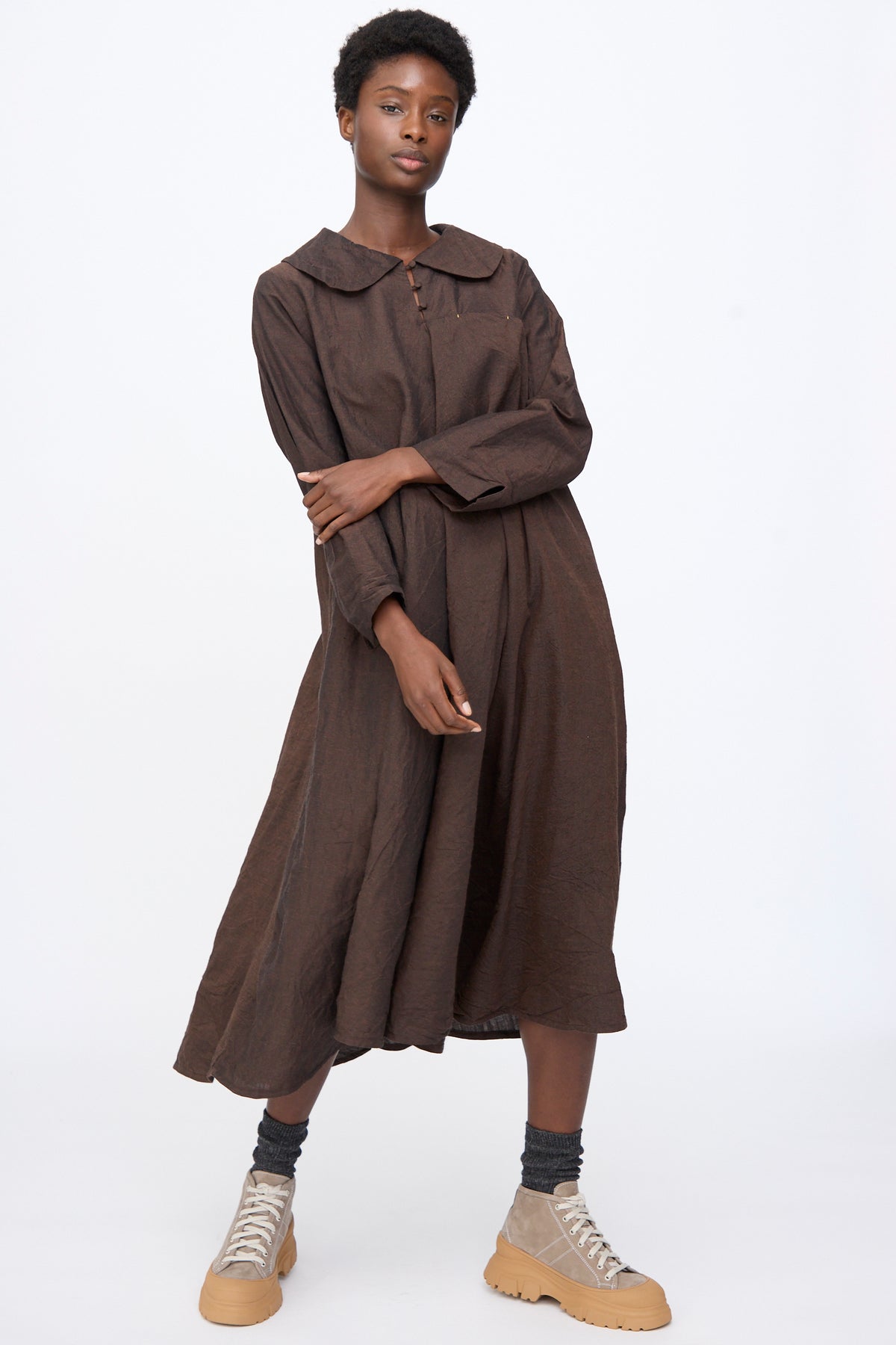 A person wearing a brown Linen Wool Robe à Col Claudine with a rounded collar, dark gray socks, and tan lug-sole sneakers.