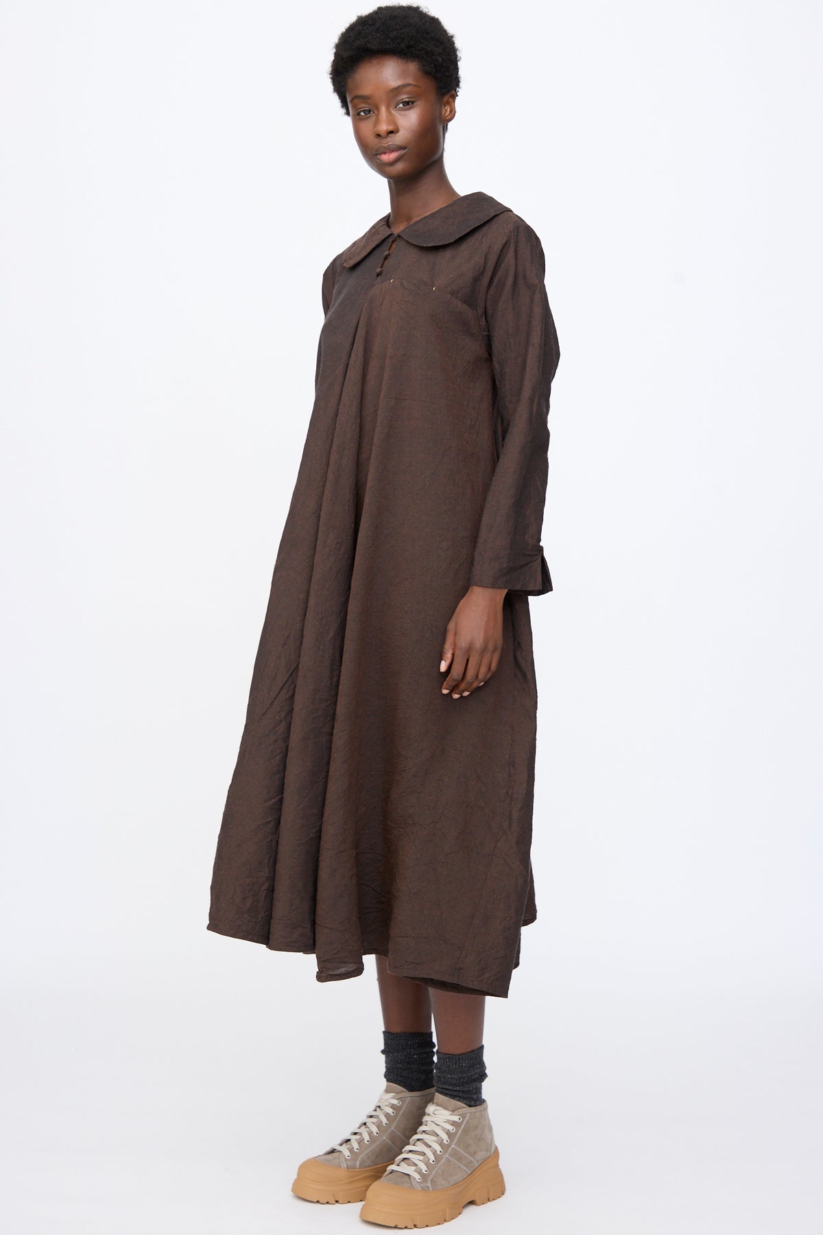 A person wearing a brown Hallelujah Linen Wool Robe à Col Claudine with a rounded collar, dark socks, and tan platform sneakers.