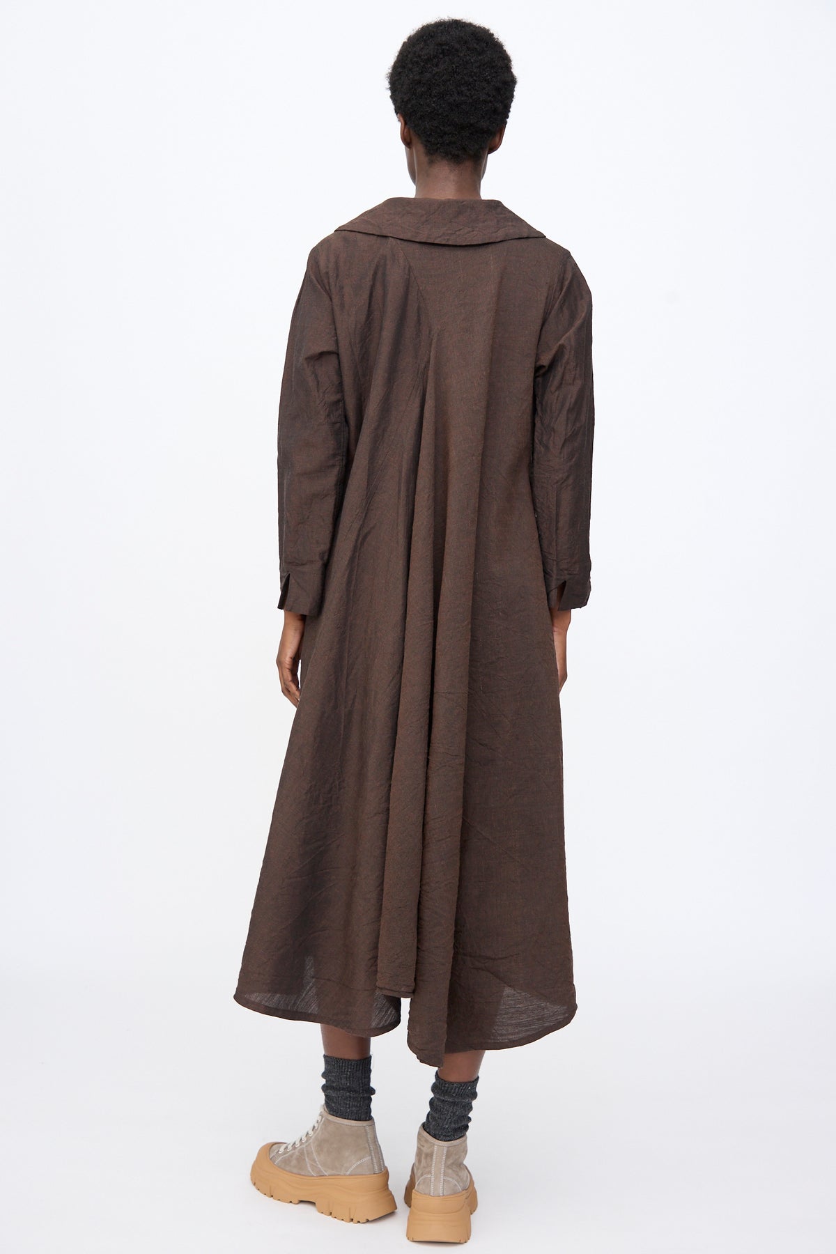 Back view of a person wearing a brown Hallelujah Linen Wool Robe à Col Claudine midi-length dress with a wide collar, grey socks, and tan platform sneakers.