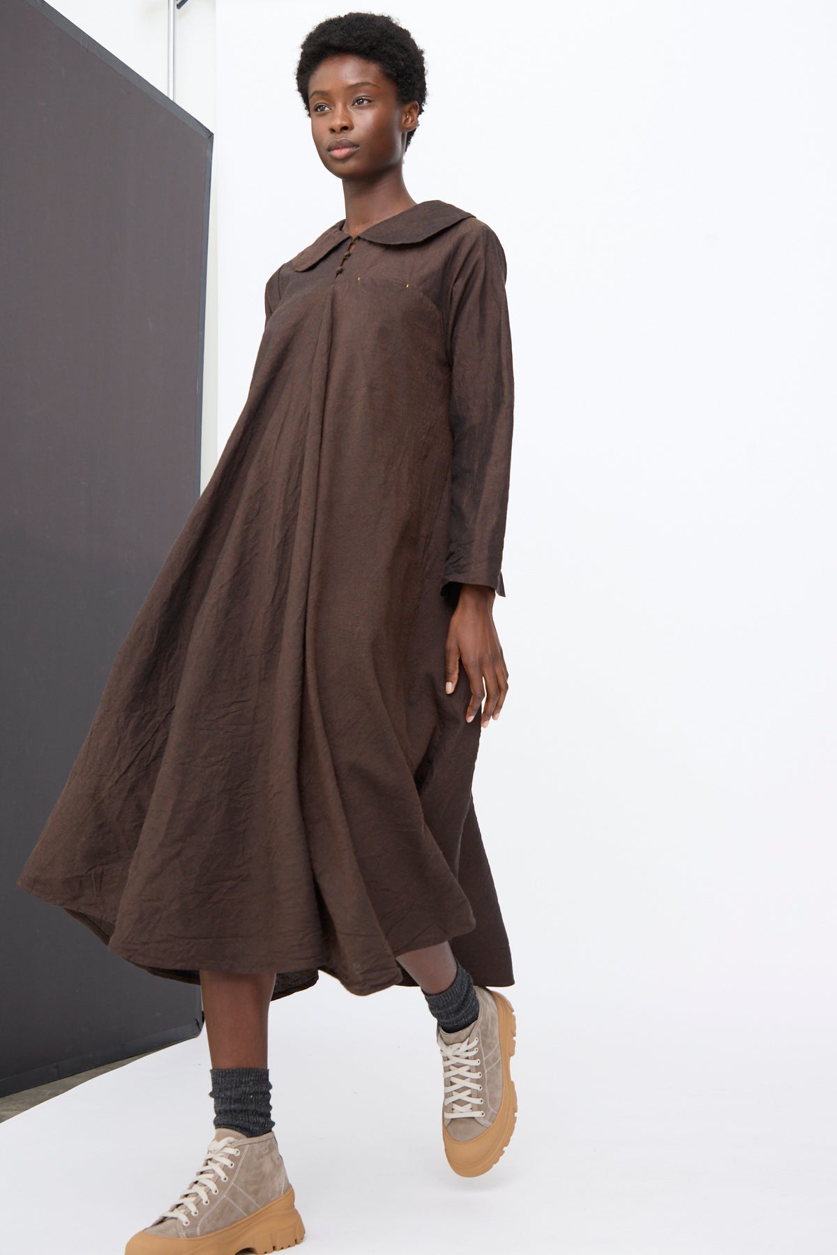 A person wears a brown Hallelujah Linen Wool Robe à Col Claudine, a long-sleeved midi dress with a rounded Peter Pan collar, paired with grey socks and tan sneakers with thick rubber soles.