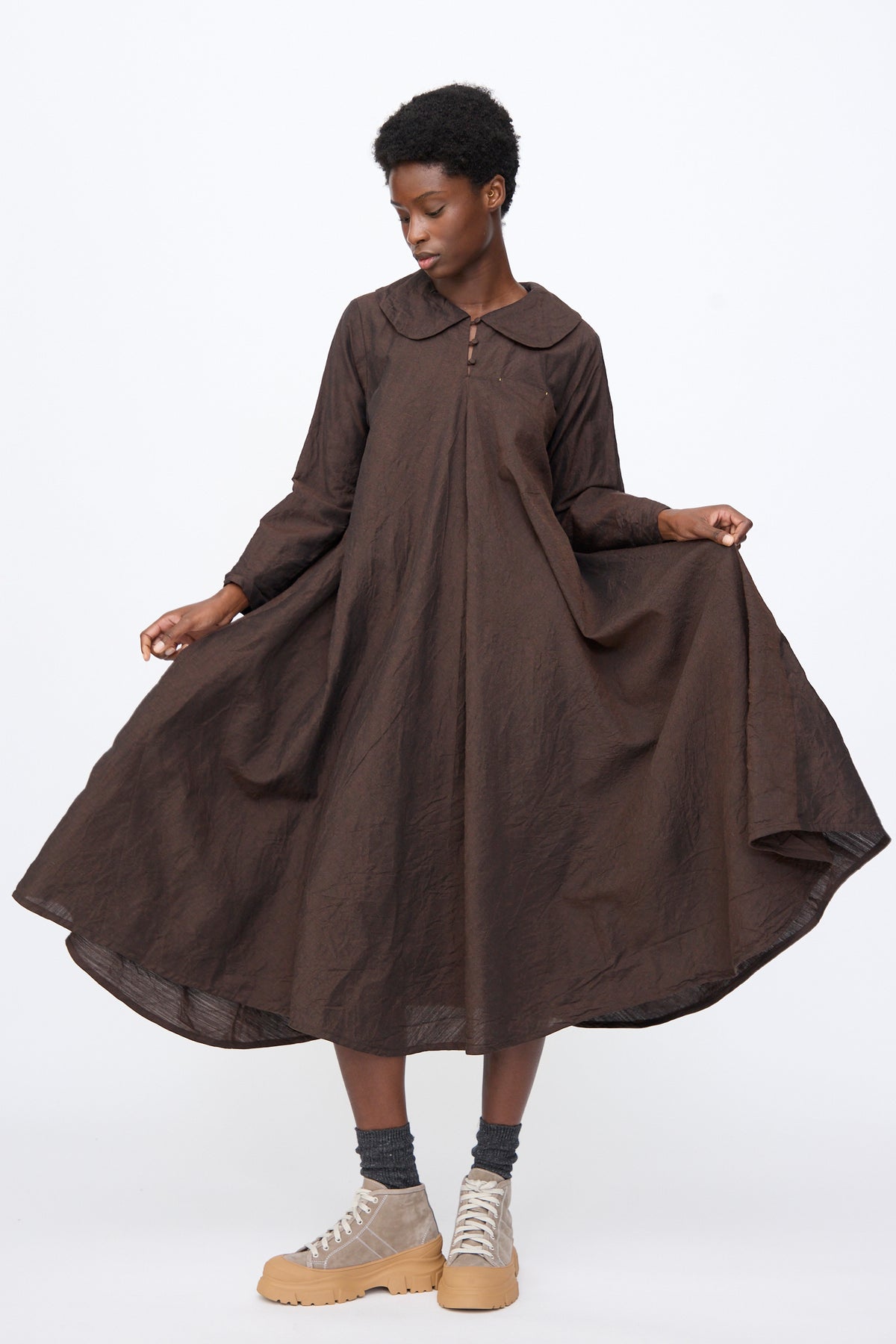 A person wearing a brown Hallelujah Linen Wool Robe à Col Claudine long-sleeved midi dress with a Peter Pan collar, grey socks, and tan high-top sneakers, holding out the full skirt.