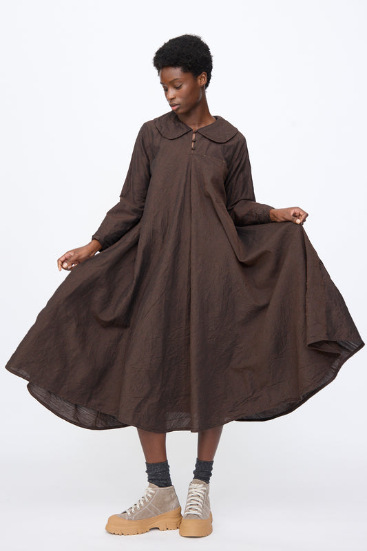 A person wearing a brown Hallelujah Linen Wool Robe à Col Claudine long-sleeved midi dress with a Peter Pan collar, grey socks, and tan high-top sneakers, holding out the full skirt.