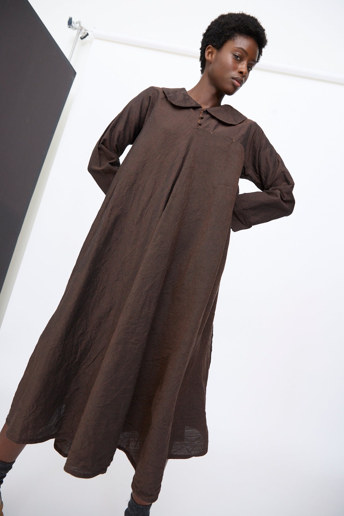 A person wearing a Hallelujah Linen Wool Robe à Col Claudine in brown, a long-sleeved, loose-fitting dress in a linen wool blend finished with a rounded Peter Pan collar.