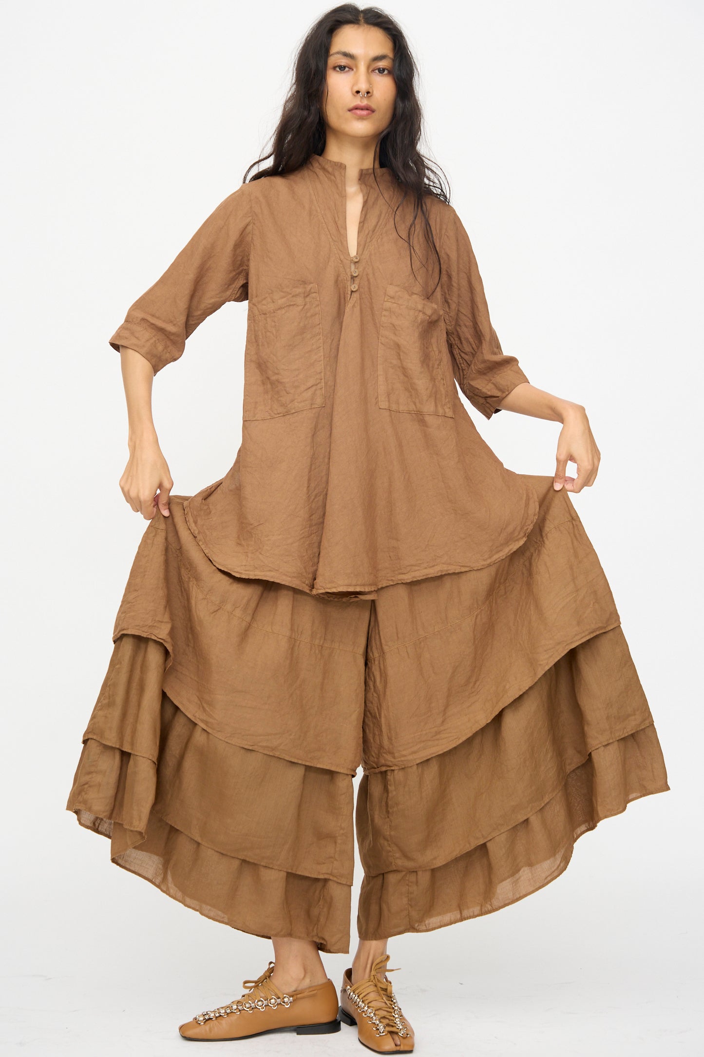 A woman stands against a white background wearing the Hallelujah Pantalon Etagée in Persimmon Dye, a loose, wide-leg linen ramie pant paired with a matching long-sleeved tunic and tan lace-up shoes.