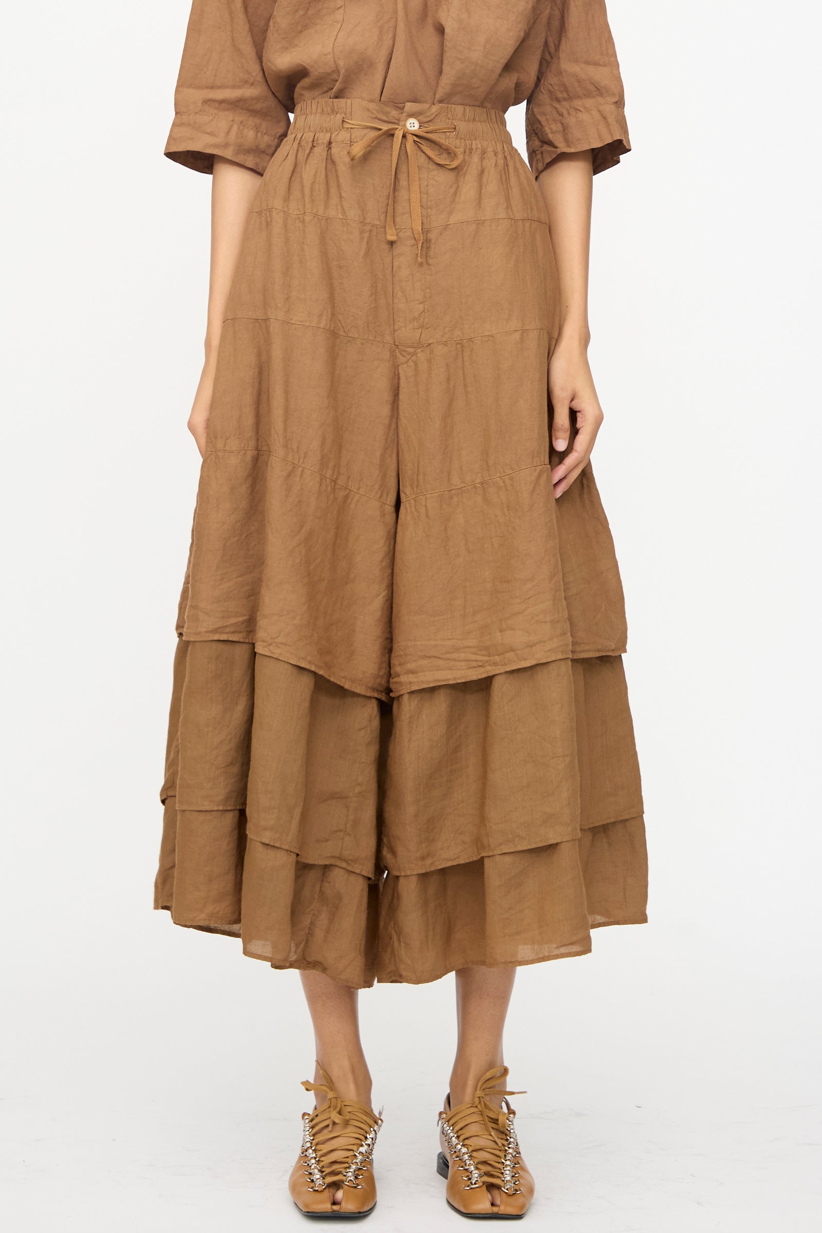 A person wears the Hallelujah Pantalon Etagée in Persimmon Dye—layered, wide-leg with a drawstring waist and cropped silhouette—paired with brown sandals, standing against a plain white background.