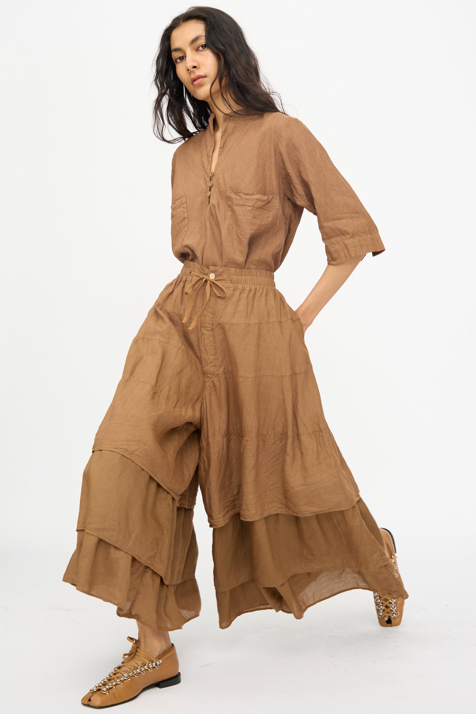 A person wears the Hallelujah Pantalon Etagée in Persimmon Dye—a loose-fitting, brown, linen-ramie button-up top and matching wide-leg pants—paired with metallic-accented flats, posing against a plain white background.
