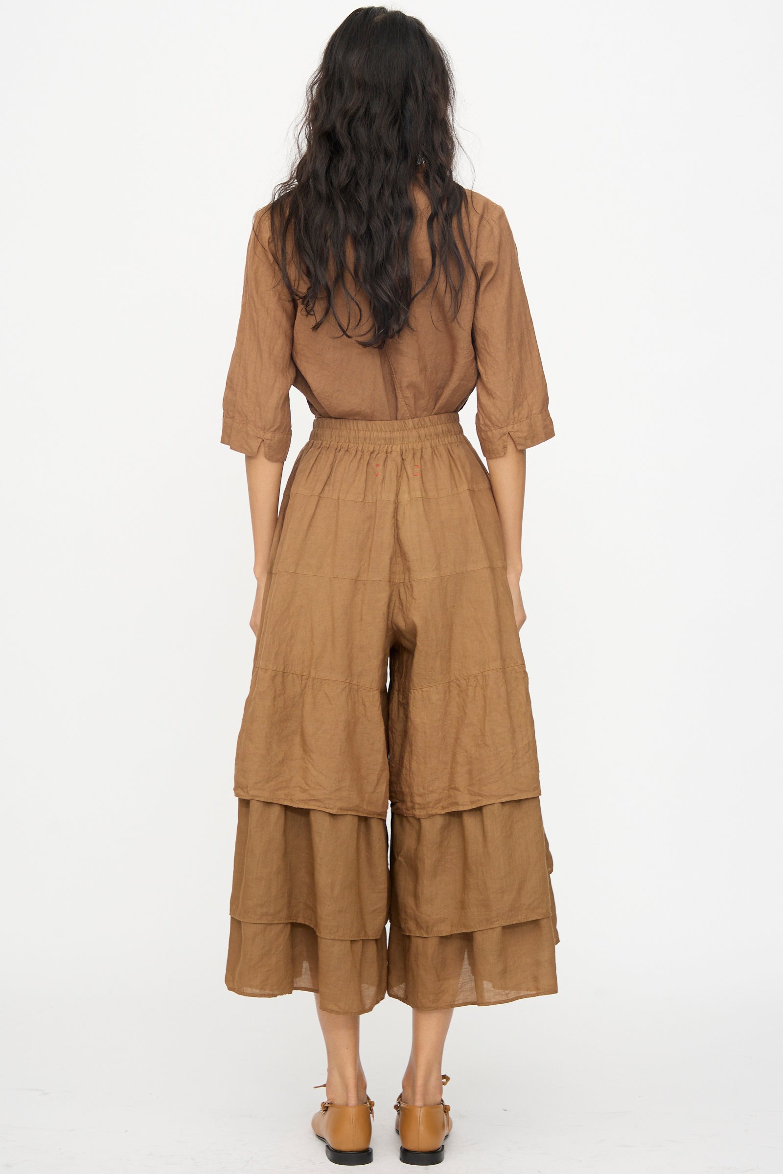 A woman with long, wavy dark hair stands facing away, wearing Hallelujah's Pantalon Etagée in Persimmon Dye and flat brown shoes.