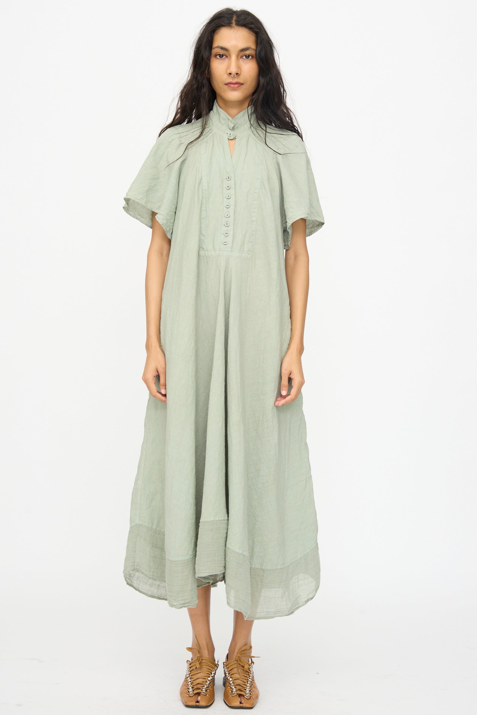 A woman faces forward wearing the Hallelujah Robe 1900’s in Green Tea Dye, a loose maxi dress with short sleeves, paired with tan woven sandals, against a plain white background.