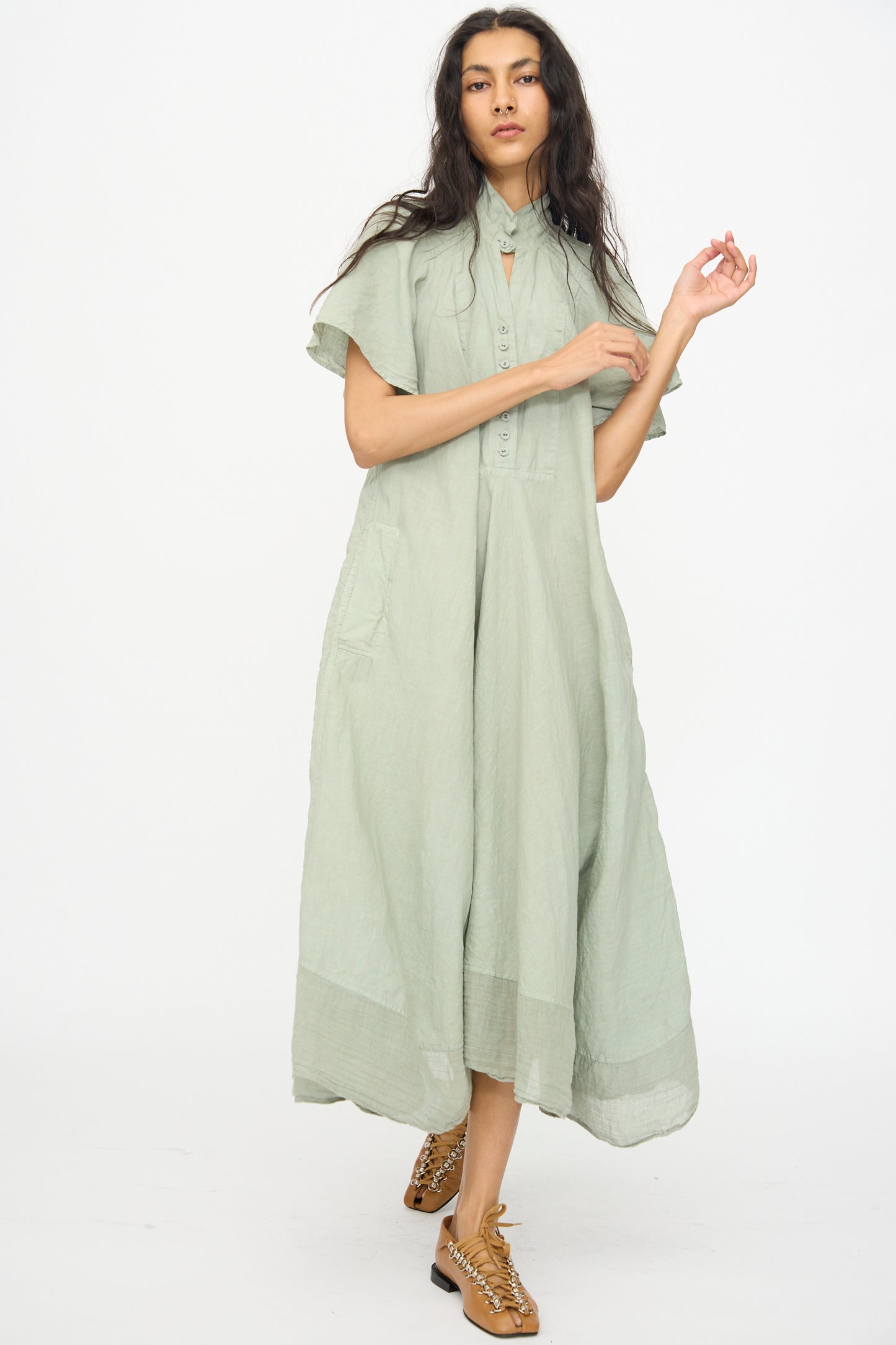 A woman stands against a white background wearing the Hallelujah Robe 1900's in Green Tea Dye, a light green, linen ramie chambray short-sleeve midi dress with pockets, paired with brown lace-up shoes.