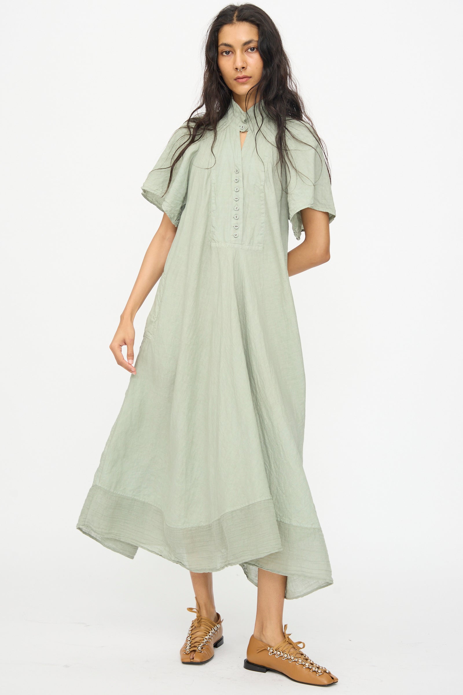 A woman wears the Hallelujah Robe 1900's in Green Tea Dye, a loose linen ramie chambray midi dress with short flutter sleeves, paired with tan flat shoes featuring decorative details, against a plain white background.