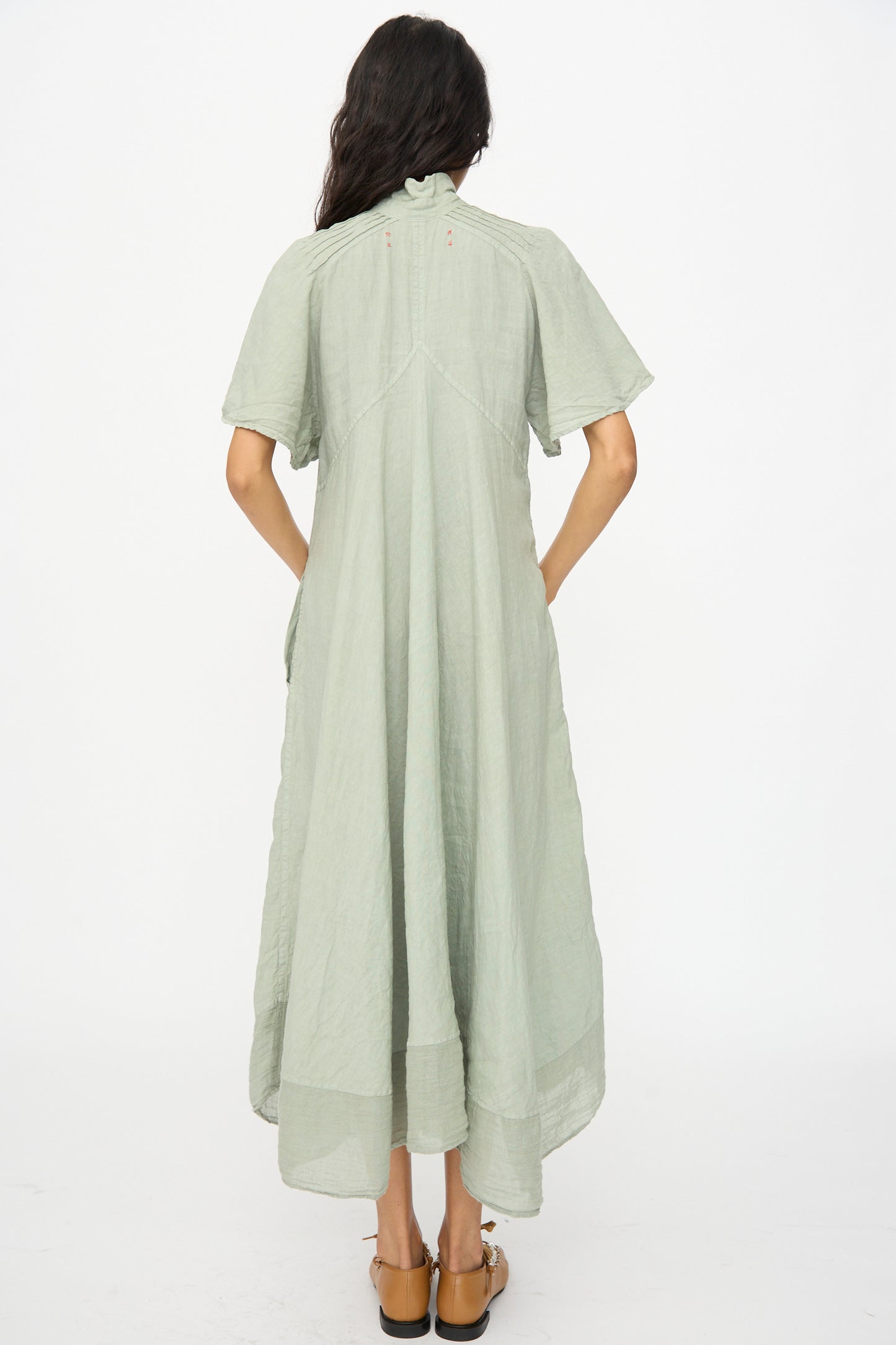 A woman stands with her back to the camera wearing the Hallelujah Robe 1900's in Green Tea Dye and brown sandals, set against a white background.